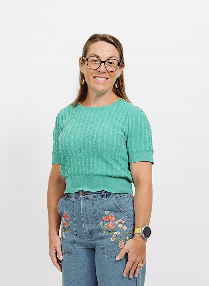 A smiling woman with glasses and long wavy hair, wearing a black t-shirt with 'Employer Branding Australia' printed on it, standing outdoors surrounded by greenery.