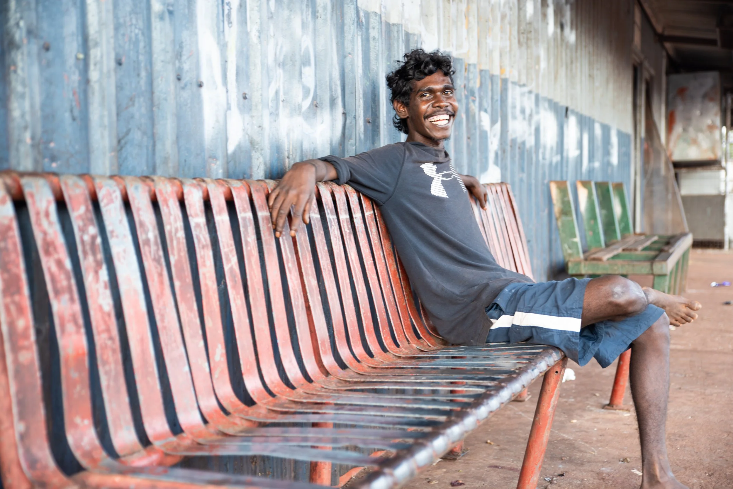 A young man with curly black hair and dark skin, smiling and sitting on a weathered red bench at an outdoor location with a corrugated metal wall behind him.