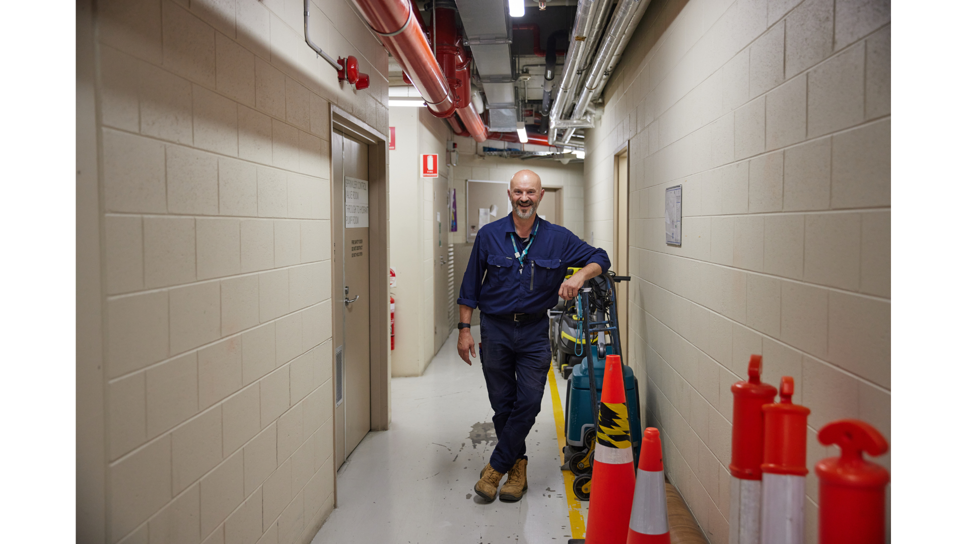 A smiling man with a beard, wearing a blue uniform, standing in a narrow hallway of an industrial or utility building. He leans on cleaning equipment like a mop and a bucket, with orange safety cones lined up along the wall in front of him. The ceiling has visible pipes and ducts, and there are fire safety signs and equipment on the walls.