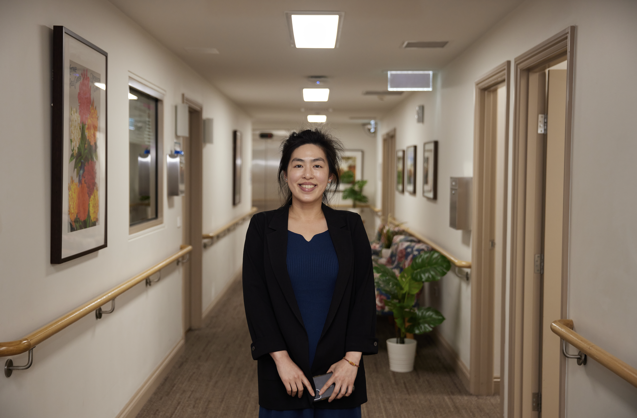 A smiling woman in a black blazer and blue top standing in a hallway with framed artwork and potted plants.