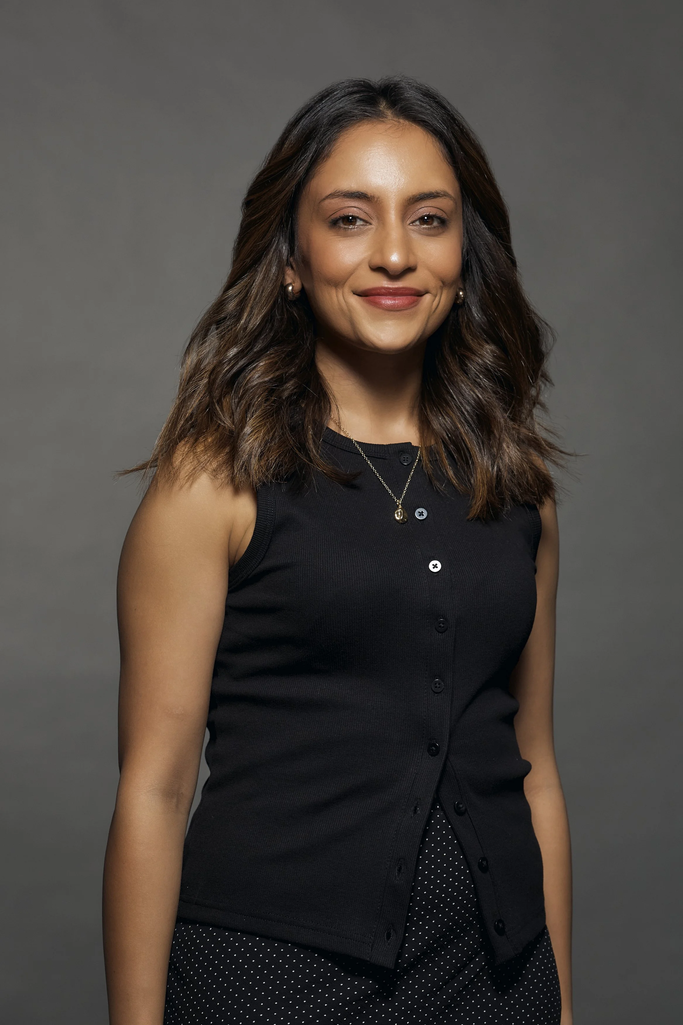 A woman with shoulder-length wavy brown hair, wearing a sleeveless black top with buttons, gold jewelry, and a polka-dot skirt, standing against a gray background with a slight smile.