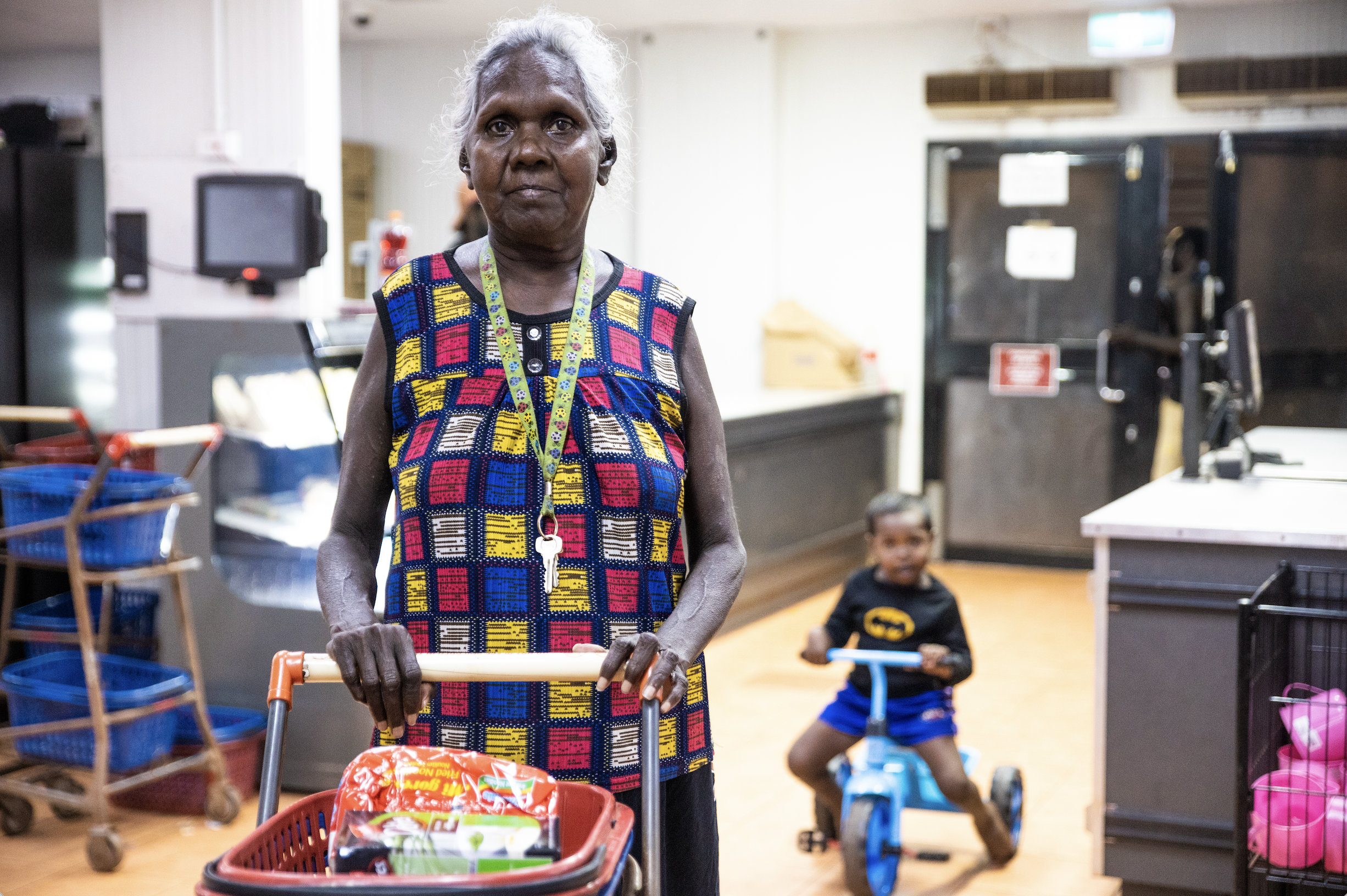 An elderly woman with gray hair and dark skin stands with a shopping cart in a store, wearing a colorful patterned sleeveless top and a lanyard with keys. A young boy with dark hair, shirtless with a Batman logo, and blue shorts rides a plastic bike in the background. The store has shelves with baskets and various items, and other shoppers are visible in the distance.