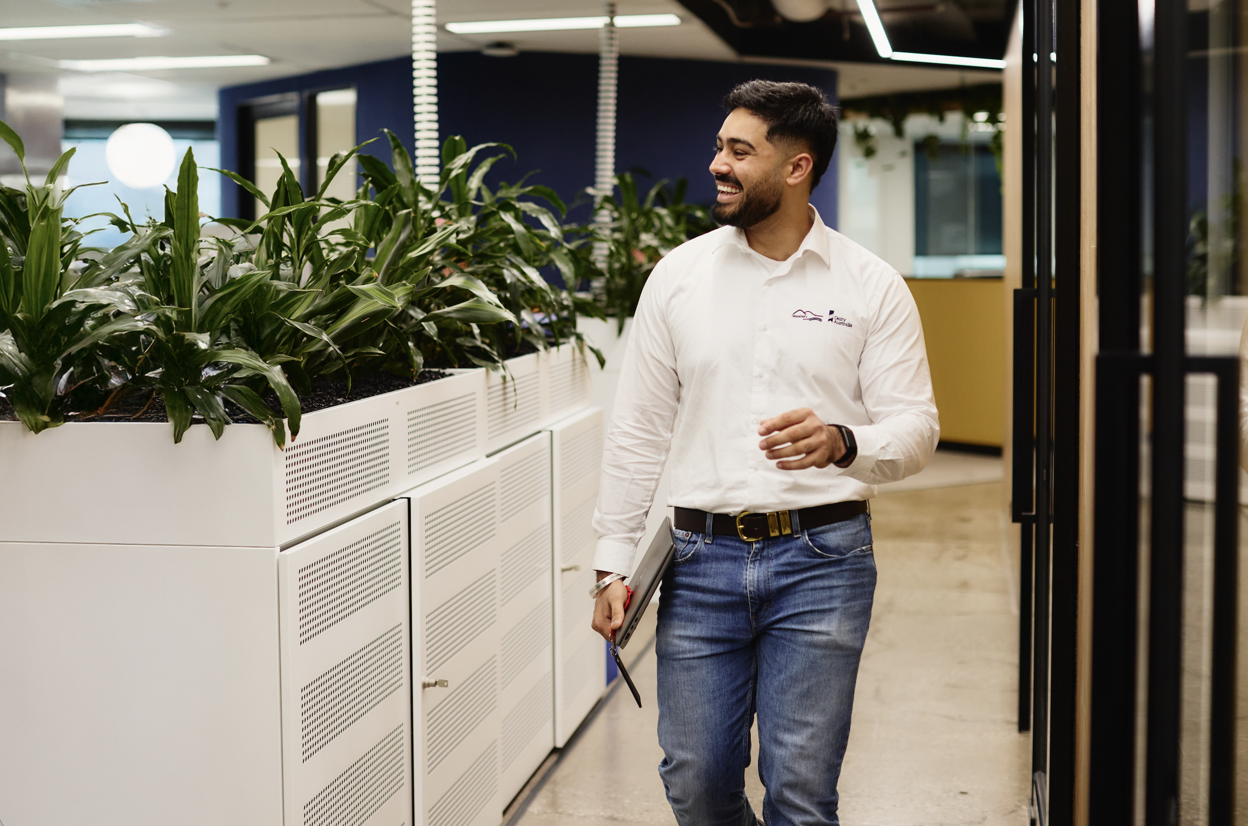 A man smiling and walking through an office or corporate environment near a row of large potted plants, holding a tablet in his left hand.
