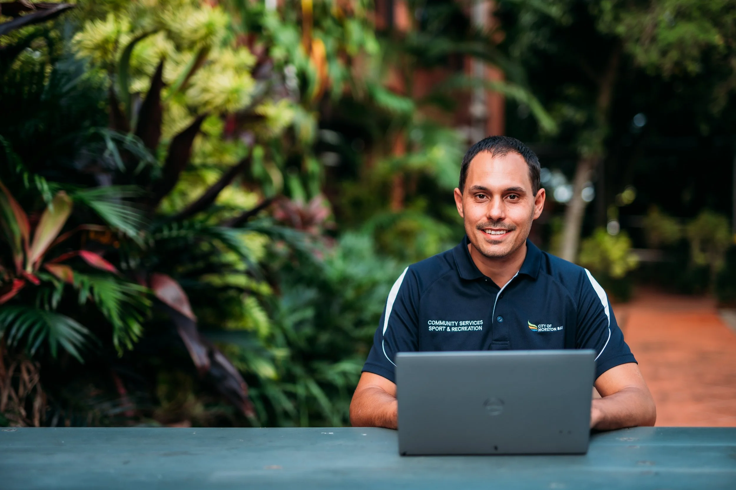 A man smiling while sitting outdoors with a laptop in front of him, surrounded by lush green plants.