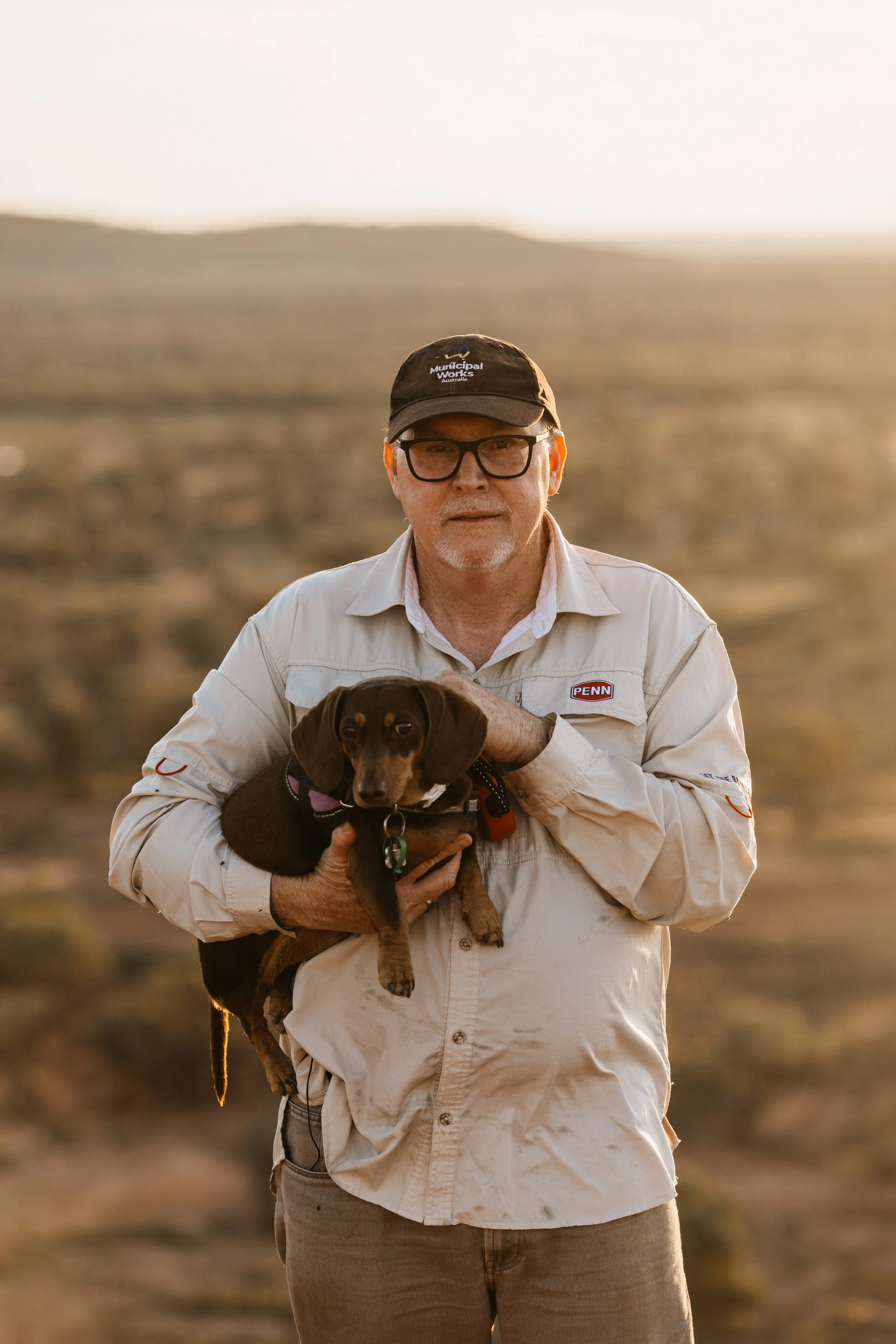 A man with glasses, a cap, and a light-colored shirt, standing outdoors in a dry, hilly landscape during sunset, holding a brown puppy.