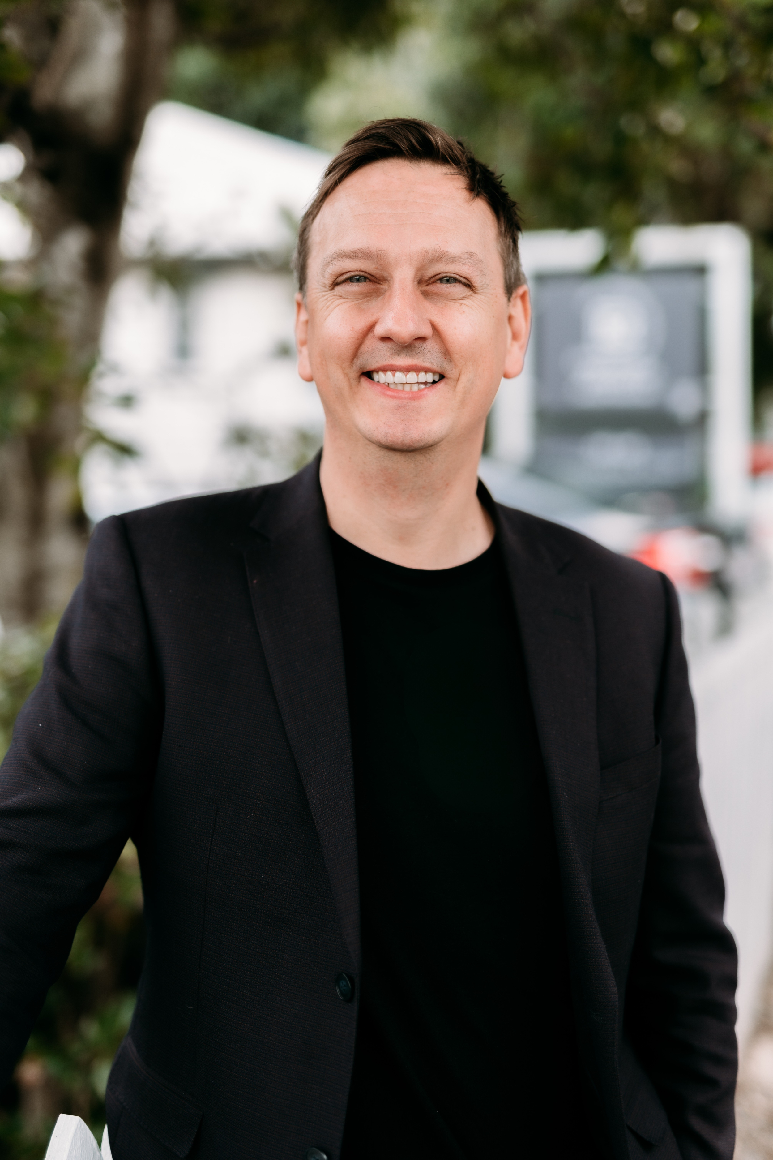 A smiling man in a black blazer and T-shirt standing outdoors with greenery and a blurred background.