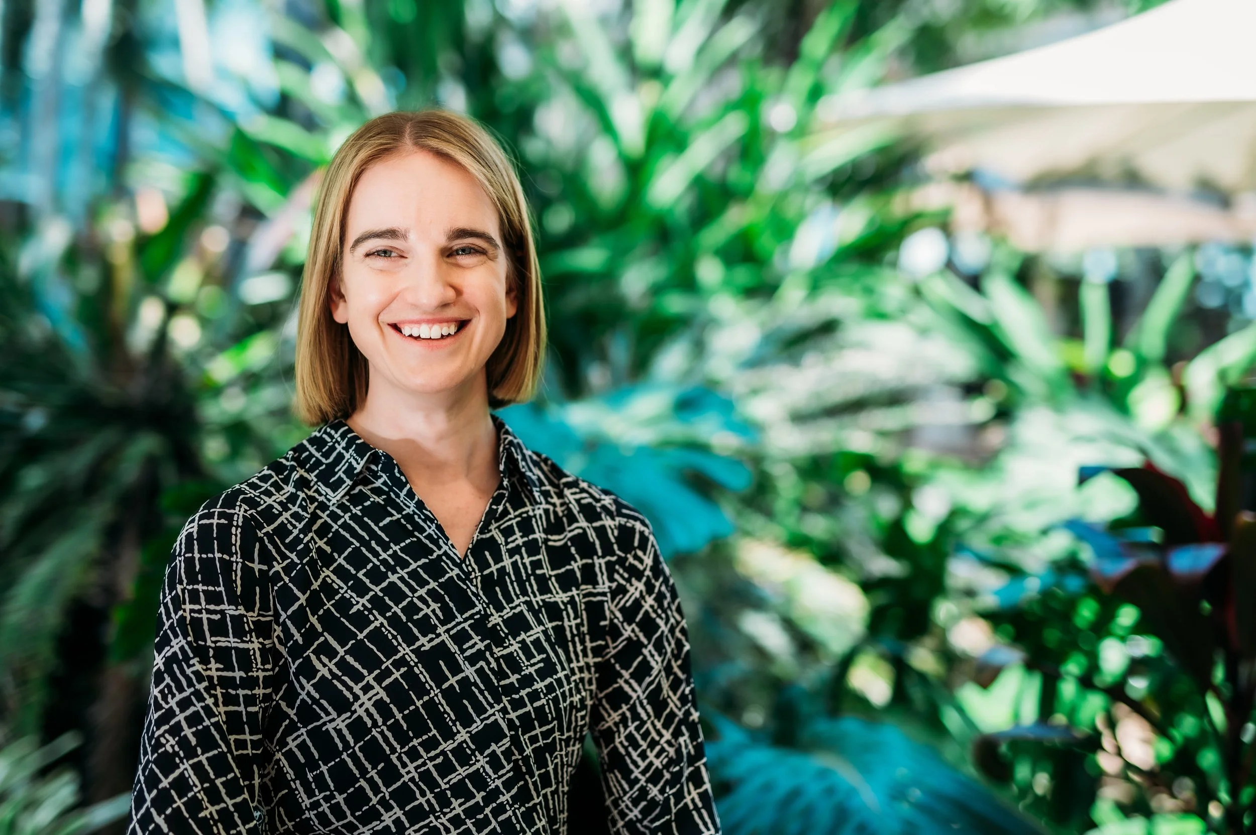A woman with shoulder-length blonde hair smiling, wearing a black and white patterned shirt, standing in front of a lush, green, tropical background.