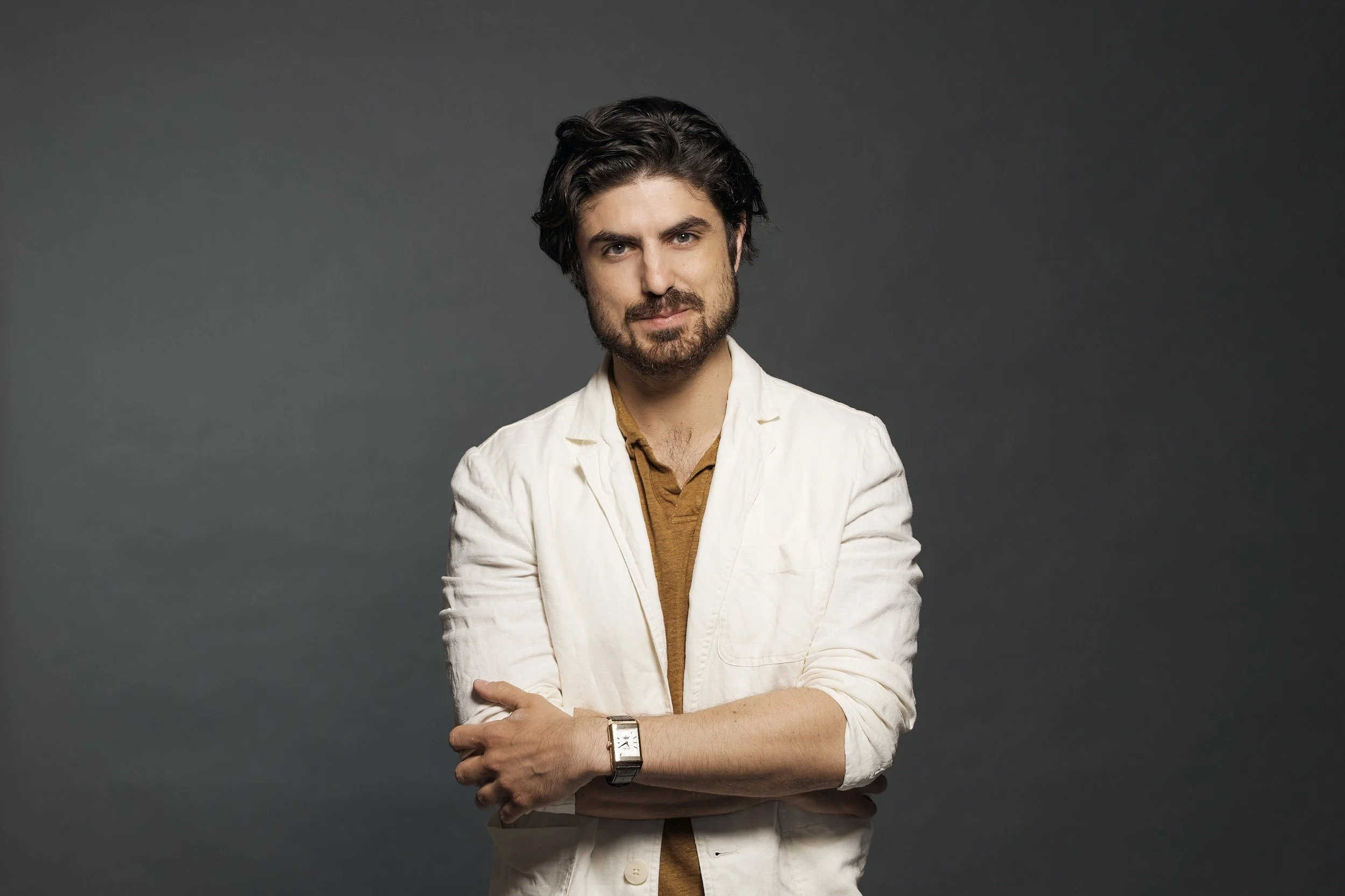 A young man with dark, wavy hair and facial hair, wearing a white blazer over a brown shirt, standing against a dark grey background with his arms crossed.