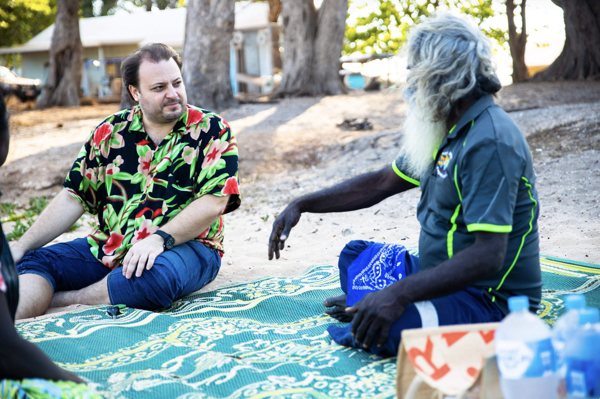 Two men sitting on a colorful beach blanket outdoors. One man has dark hair, a beard, and is wearing a colorful floral shirt. The other has gray hair, a beard, and is wearing a dark gray jacket with neon accents. They are near trees with a house and a vehicle visible in the background.