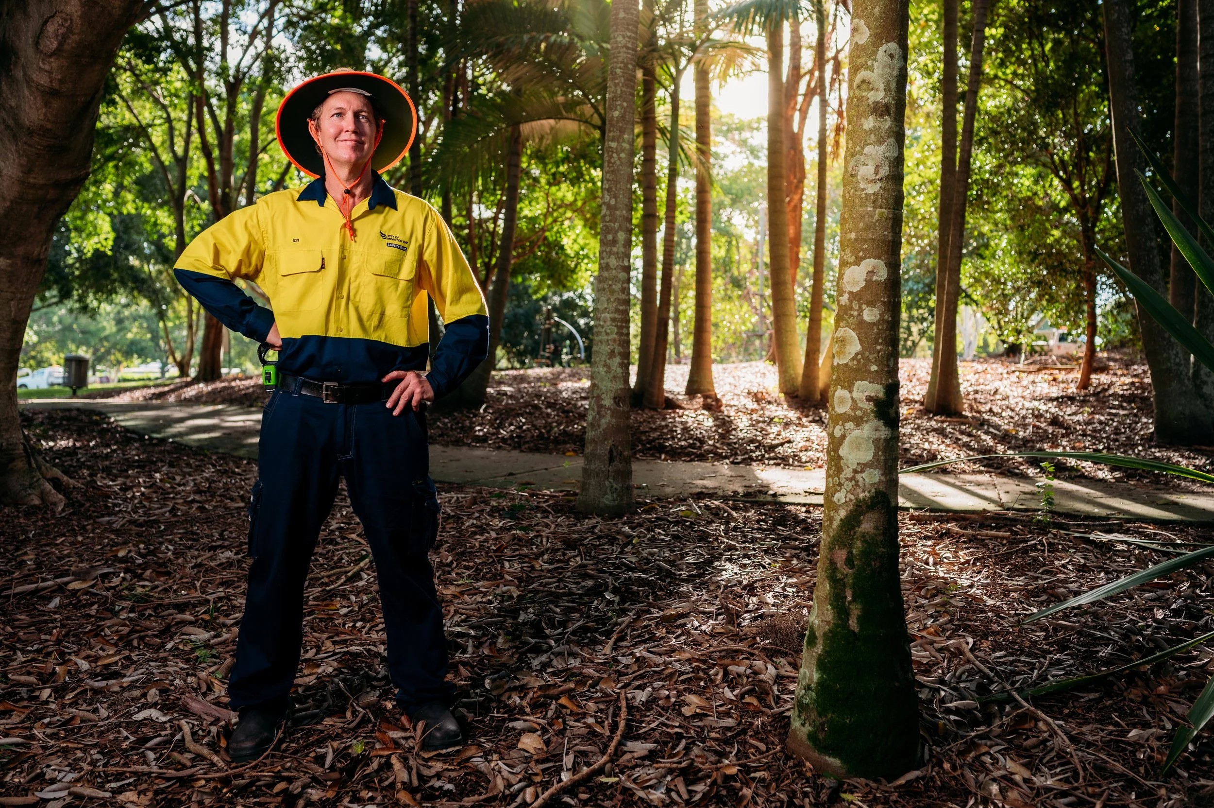A park ranger or park employee wearing a yellow jacket and wide-brimmed hat, standing on leaf-covered ground among trees, with sunlight filtering through the foliage in the background.