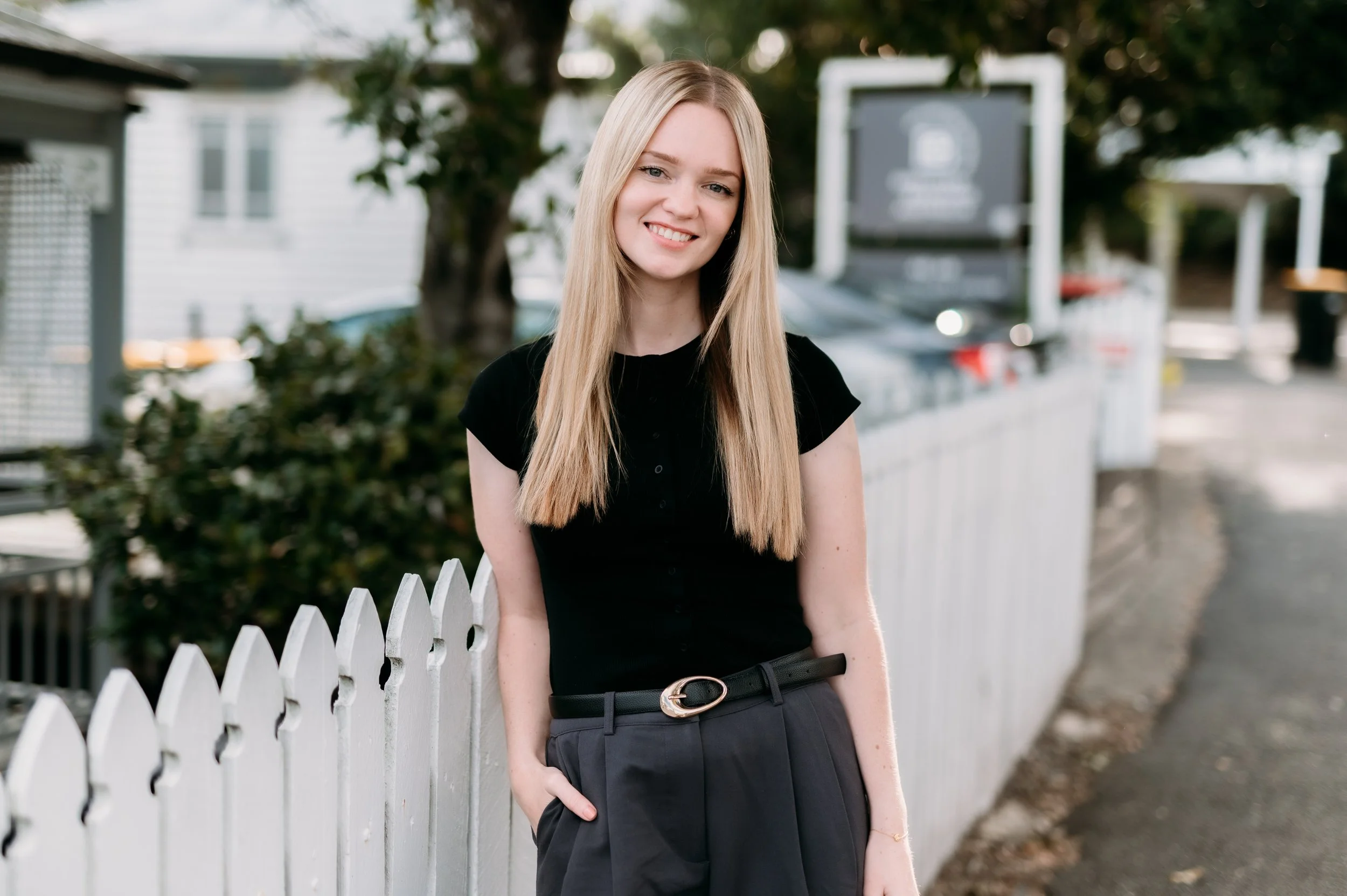A young woman with long blonde hair, wearing a black top and gray pants, posing outdoors next to a white picket fence, smiling, with trees and a building in the background.