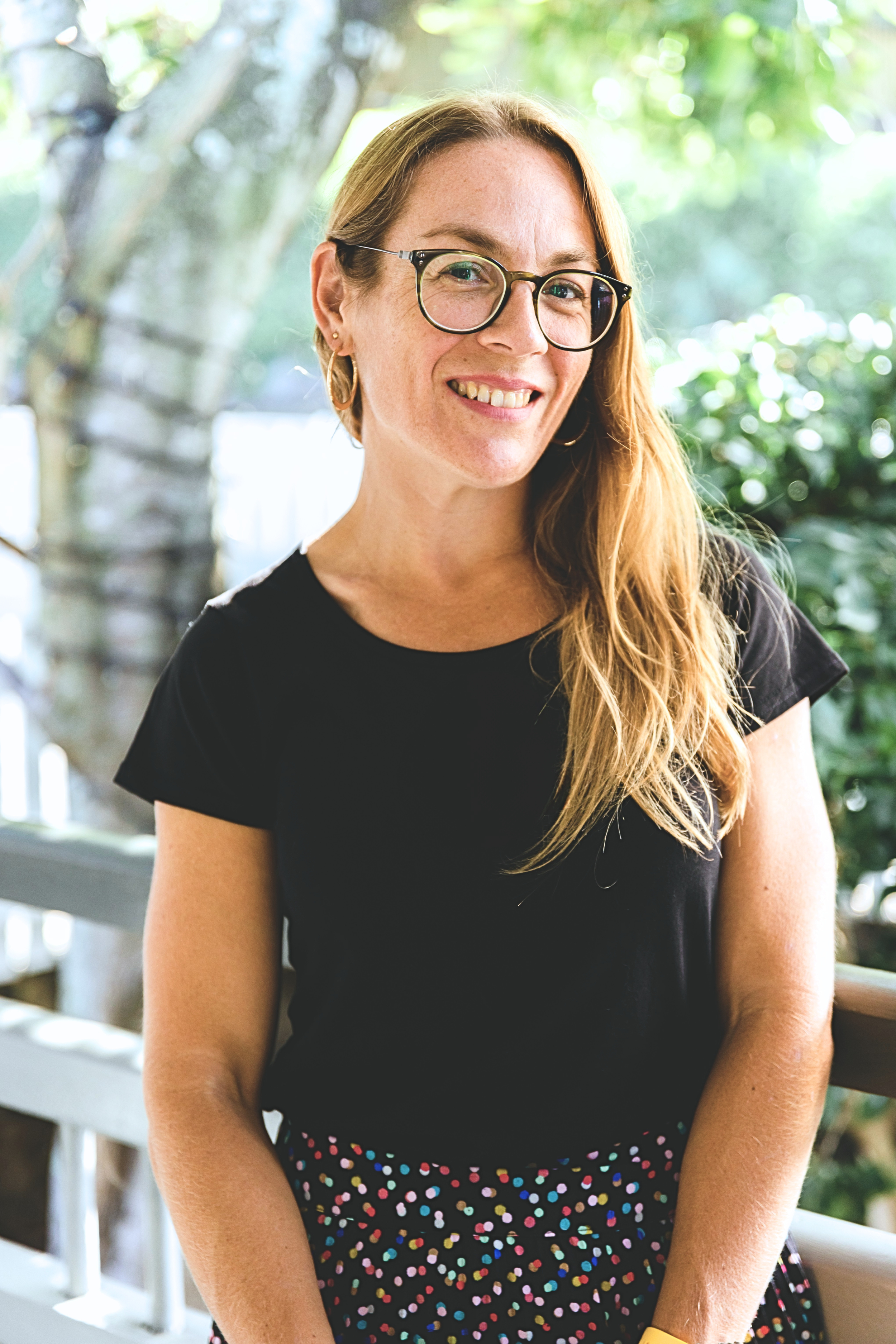 A smiling woman with glasses and long wavy hair, wearing a black t-shirt with 'Employer Branding Australia' printed on it, standing outdoors surrounded by greenery.