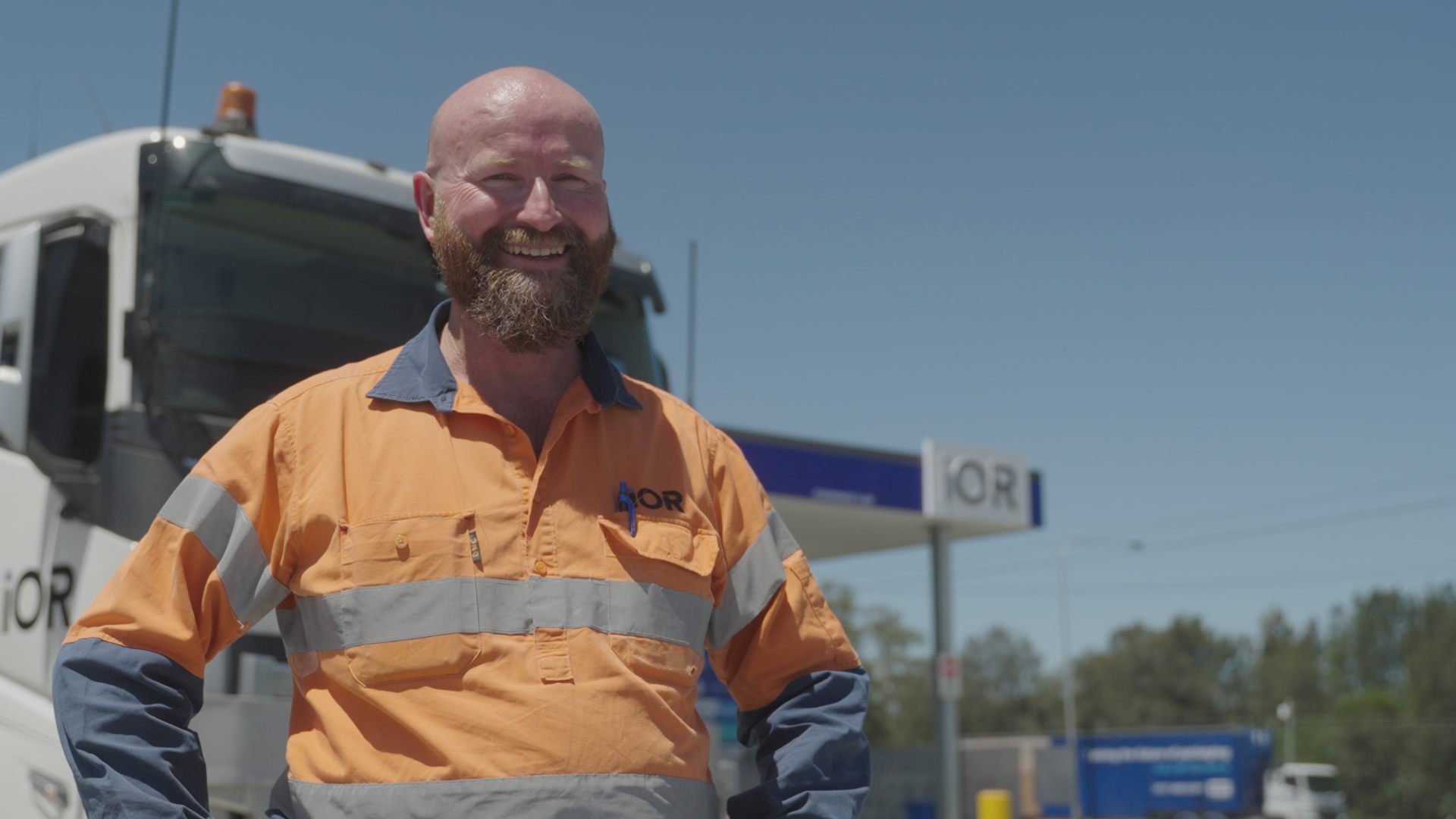 A smiling man with a beard wearing an orange safety uniform stands outdoors in front of a helicopter and a gas station sign.