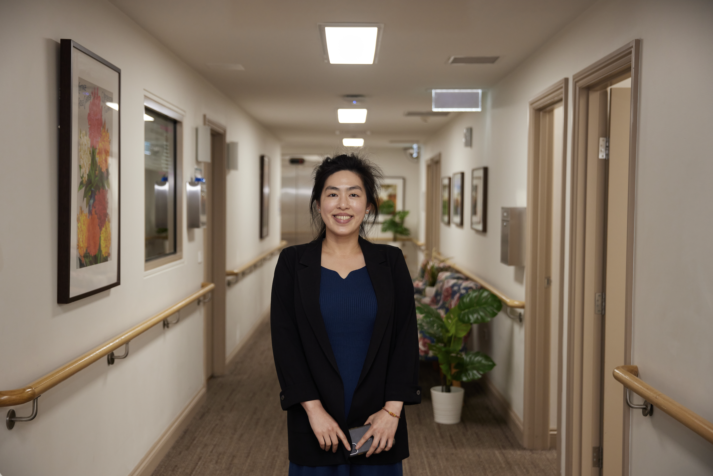 A smiling woman with dark hair standing in a hallway with beige walls, artwork on the walls, plants, and several doors.