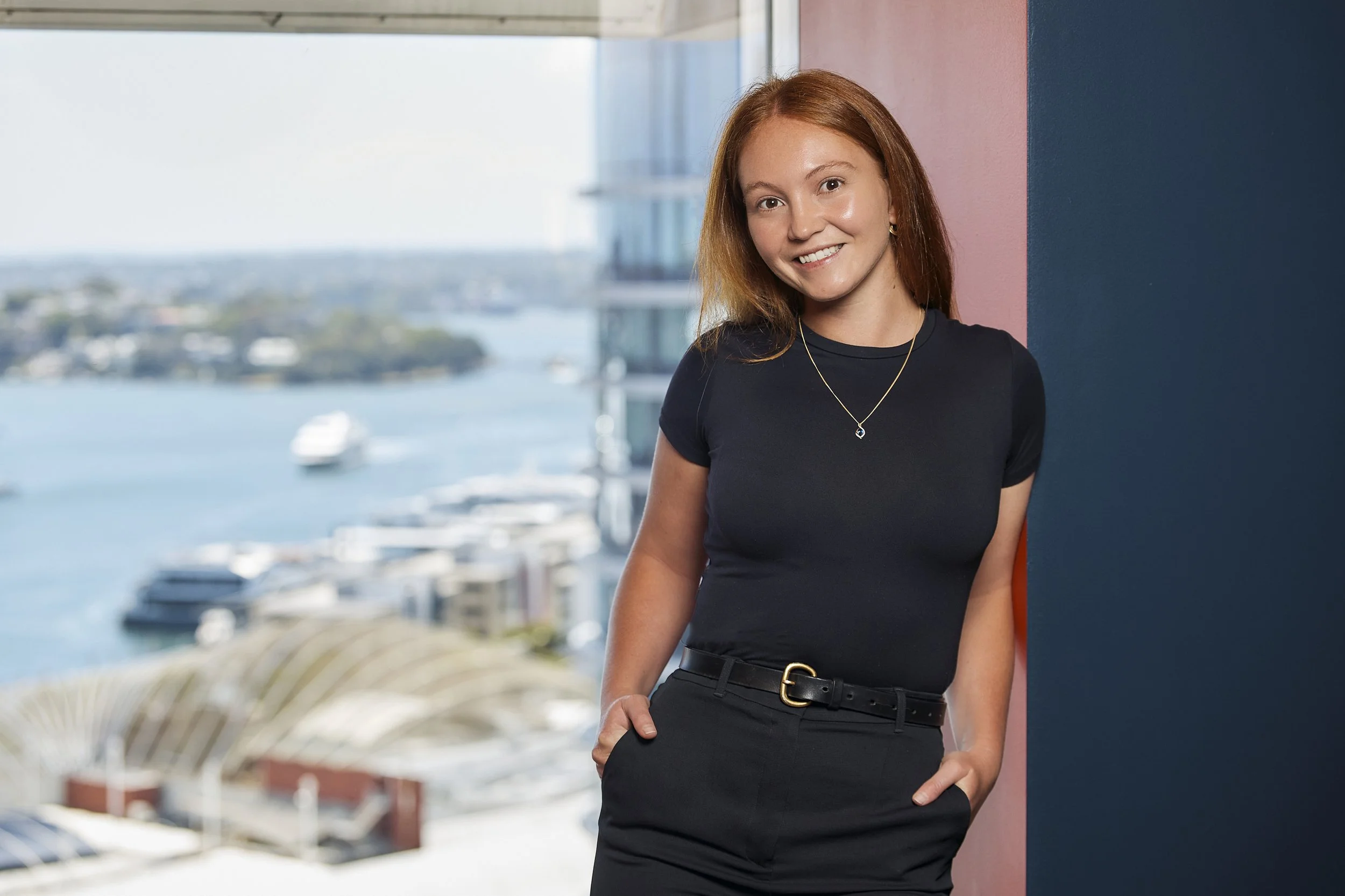 A young woman with red hair standing by a colorful wall, smiling at the camera with a harbor view in the background.