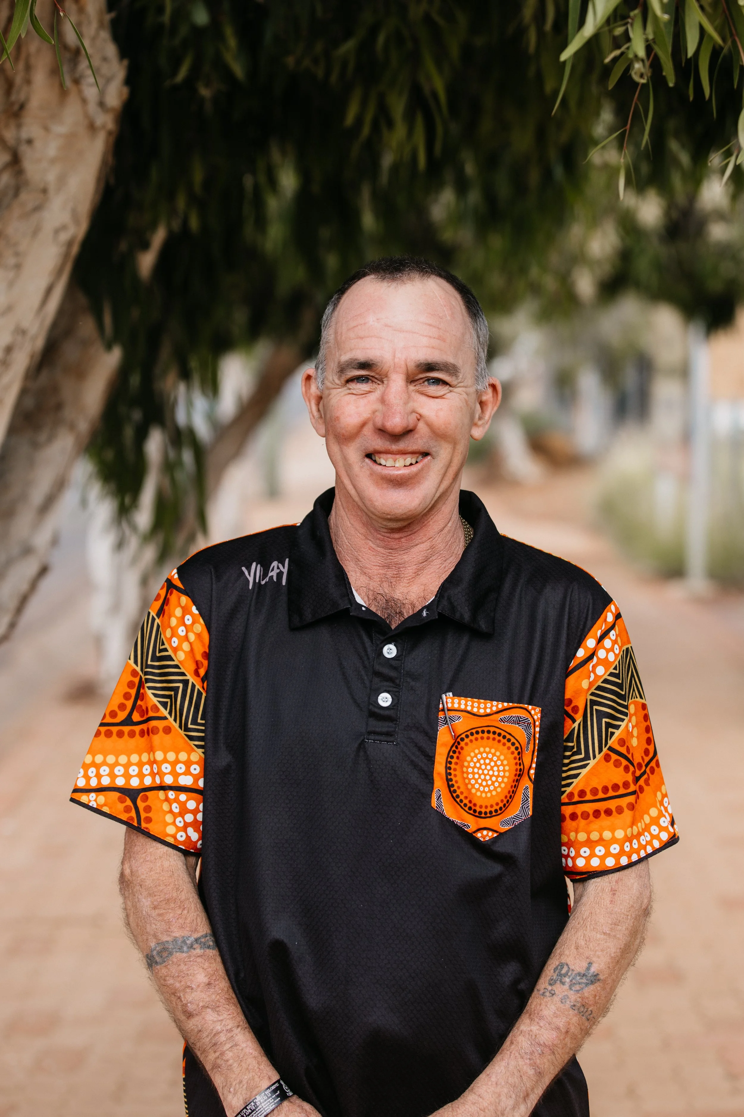 A middle-aged man with short dark hair, wearing a black shirt with orange and patterned accents on the sleeves and pocket, standing outdoors near trees and a pathway.