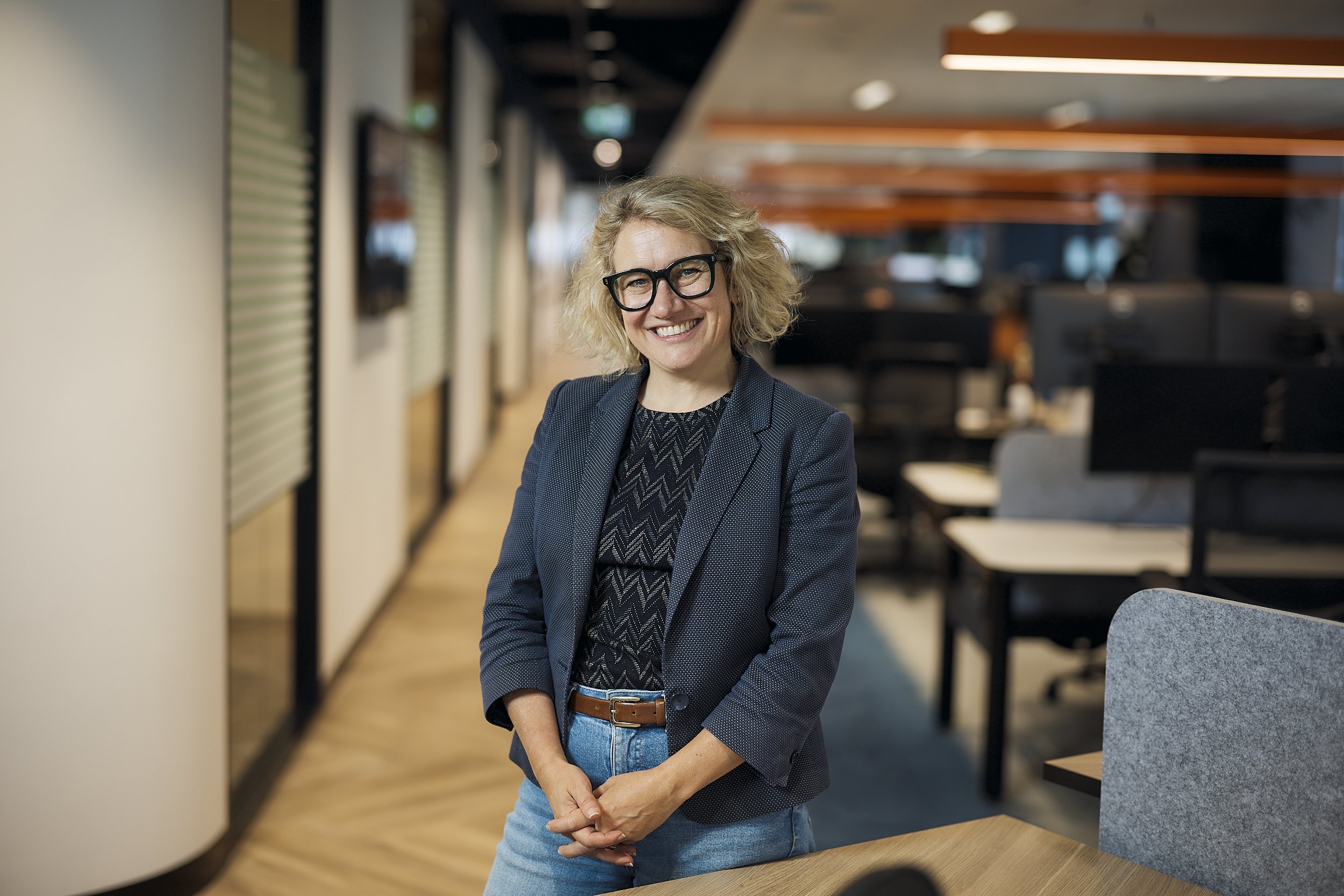 A smiling woman with curly blonde hair and glasses wearing a dark blazer and patterned top, standing in an office environment with wooden accents and multiple computer workstations in the background.