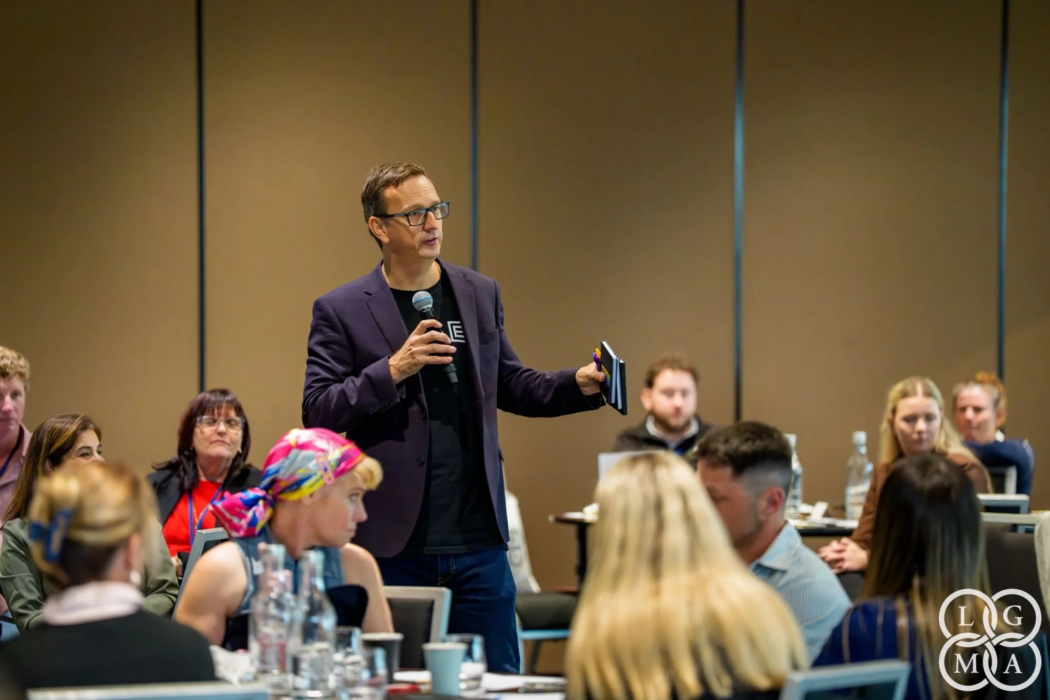 A man with glasses and a dark purple blazer is holding a microphone and speaking to an audience in a conference room. Several audience members, a mix of men and women, are seated at tables, listening attentively. The background features plain beige walls.
