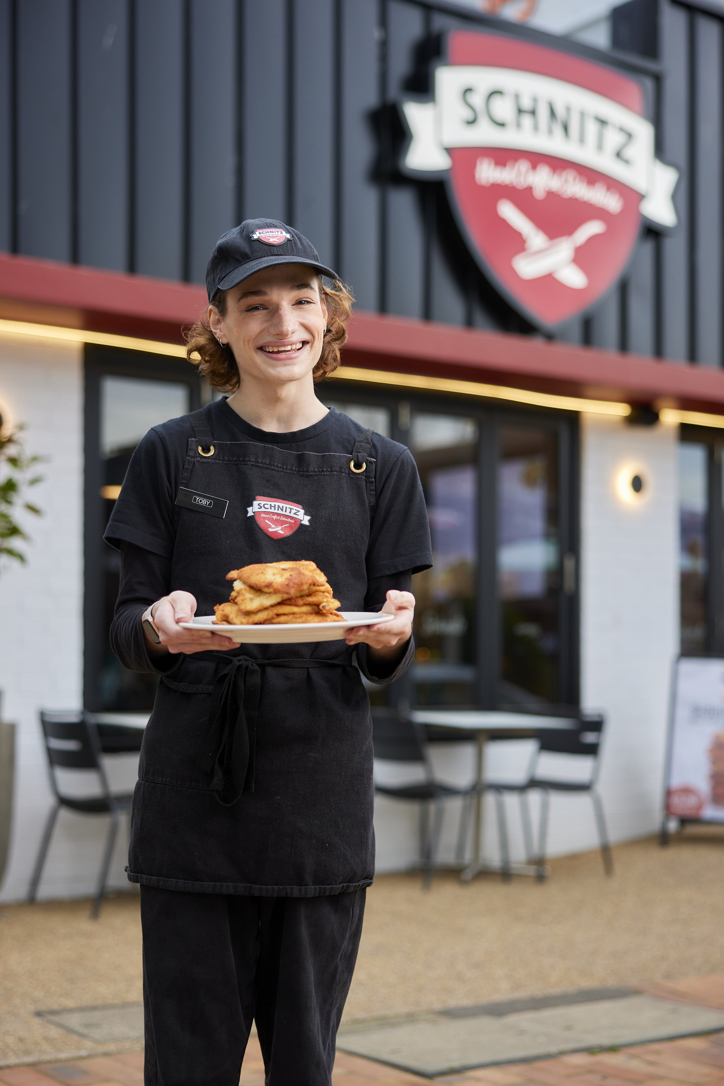 A smiling female server in a black apron and hat holding a plate of fried chicken outside a restaurant with a Schnitz sign.