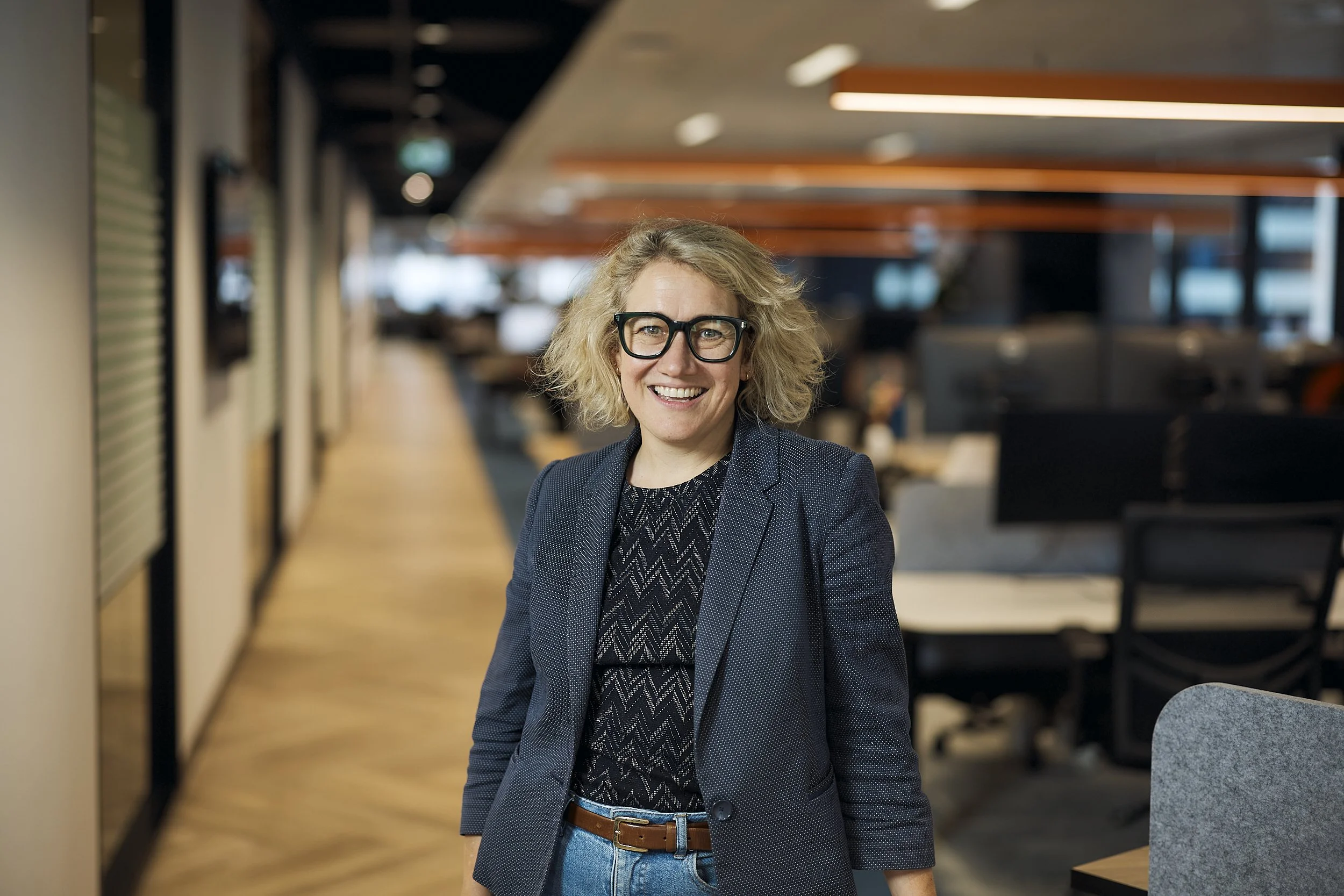 Smiling woman in black glasses, blazer, and jeans in office corridor with JavaScript posters on wall.