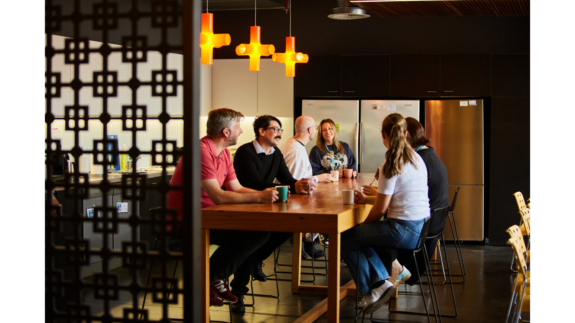 A group of six people sit around a wooden table in a modern, stylish kitchen or cafeteria. They are engaged in conversation, smiling, and holding mugs. The space features black cabinets, two stainless steel refrigerators, and warm orange pendant lights hanging from the ceiling.