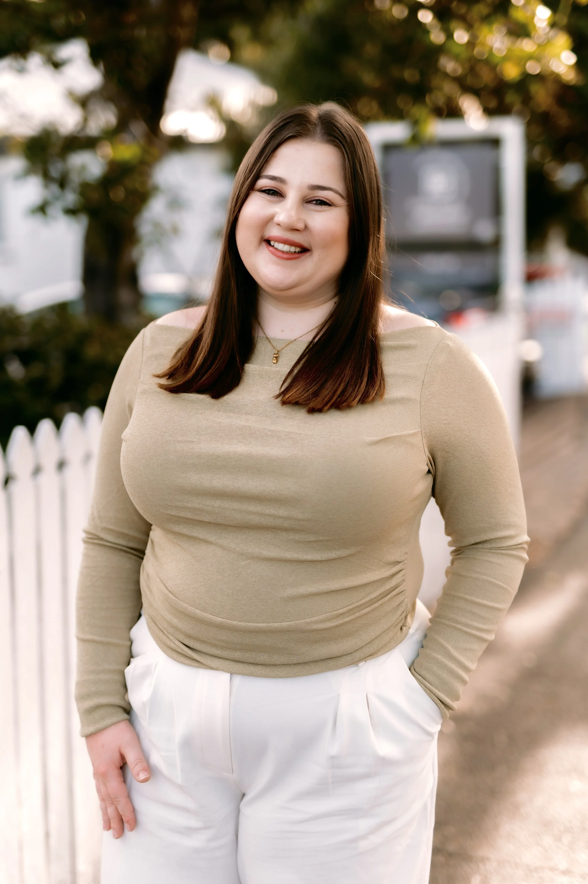 A woman with shoulder-length brown hair smiling outdoors, wearing a beige long-sleeve top and white pants, standing near a white picket fence with trees in the background.