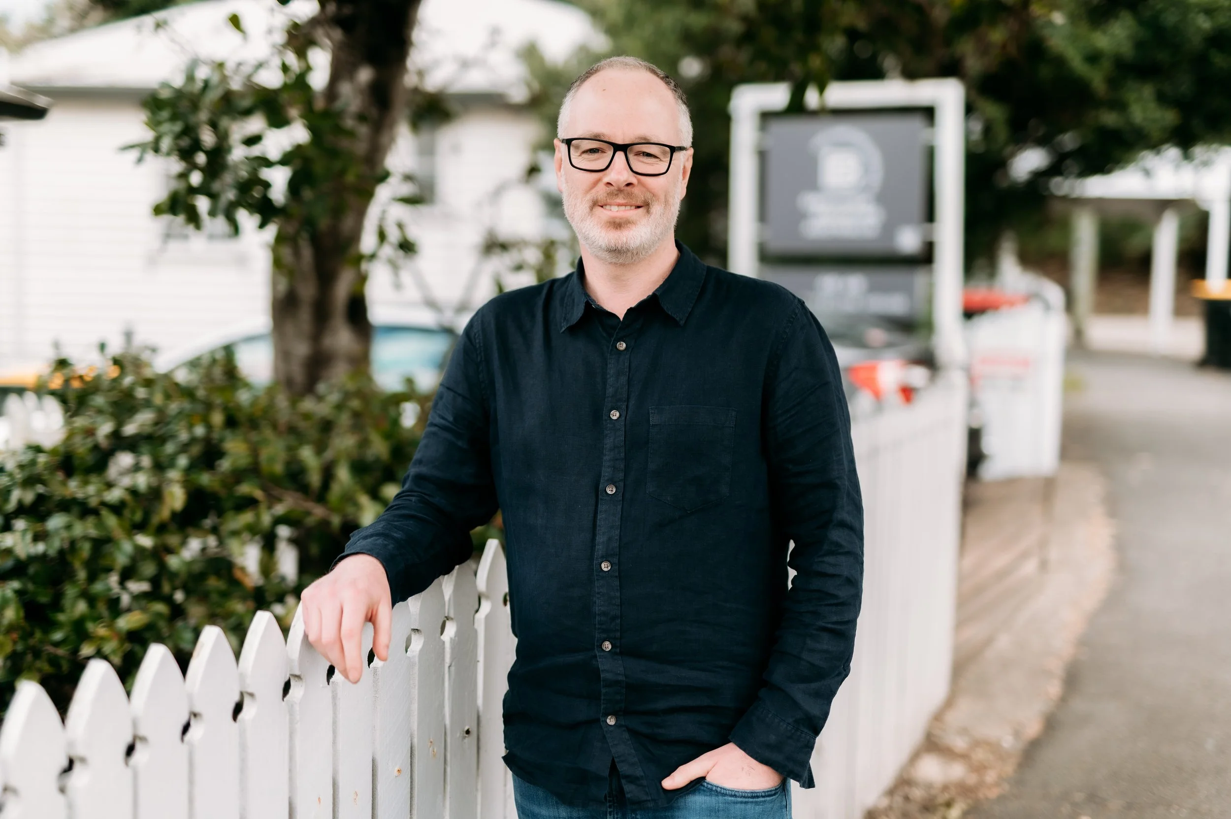 Man with glasses, gray beard, wearing a black shirt, standing outside near a white fence.