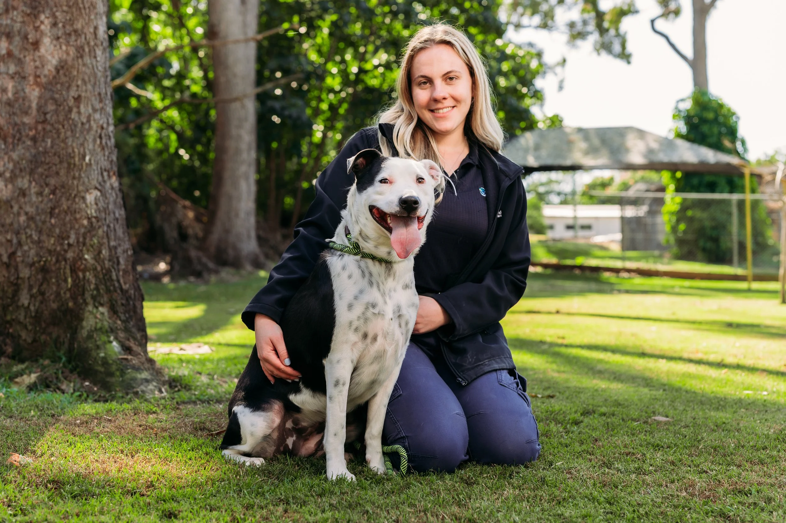A woman with blonde hair kneeling on the grass outdoors, smiling, with a black and white dog sitting beside her. The background includes trees, a fence, and a shaded picnic area.