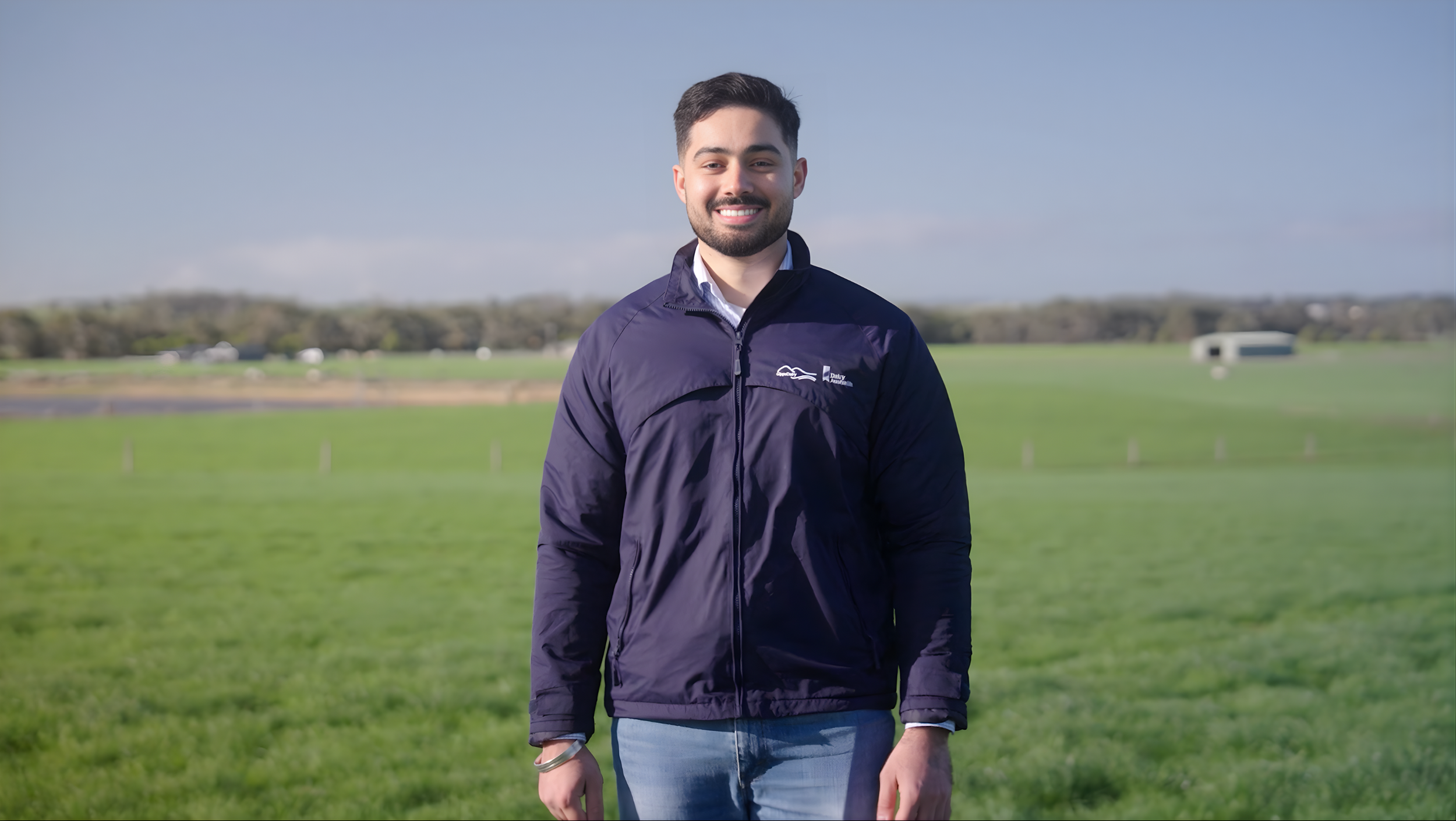 A smiling young man with dark hair and a beard standing outdoors in a field with a clear blue sky in the background, wearing a dark jacket and light jeans.
