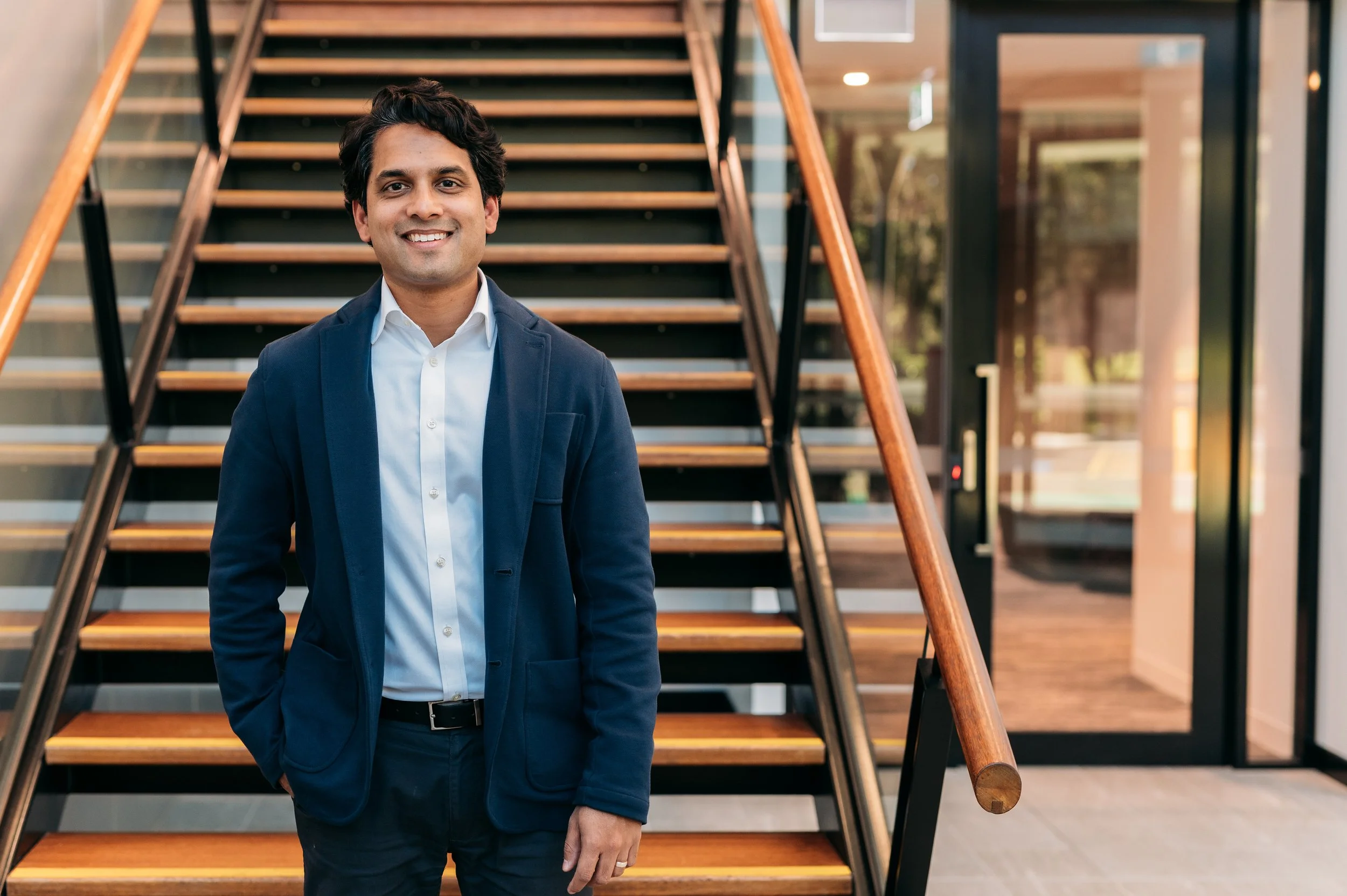 A smiling man in a navy blazer and white shirt standing in front of a staircase inside a modern building with glass walls.