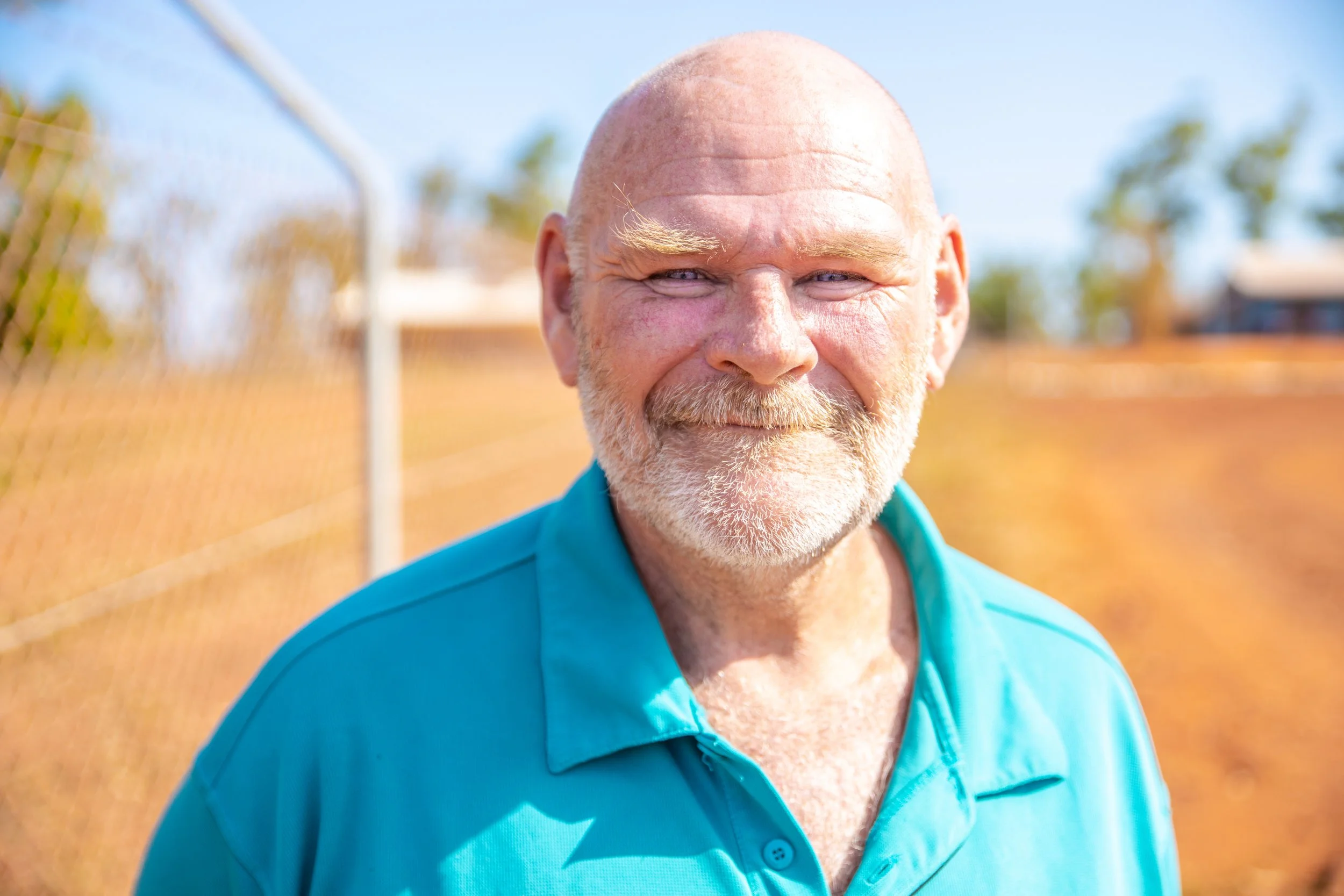 Smiling older man with a bald head and beard wearing a bright blue polo shirt, standing outdoors near a chain-link fence.