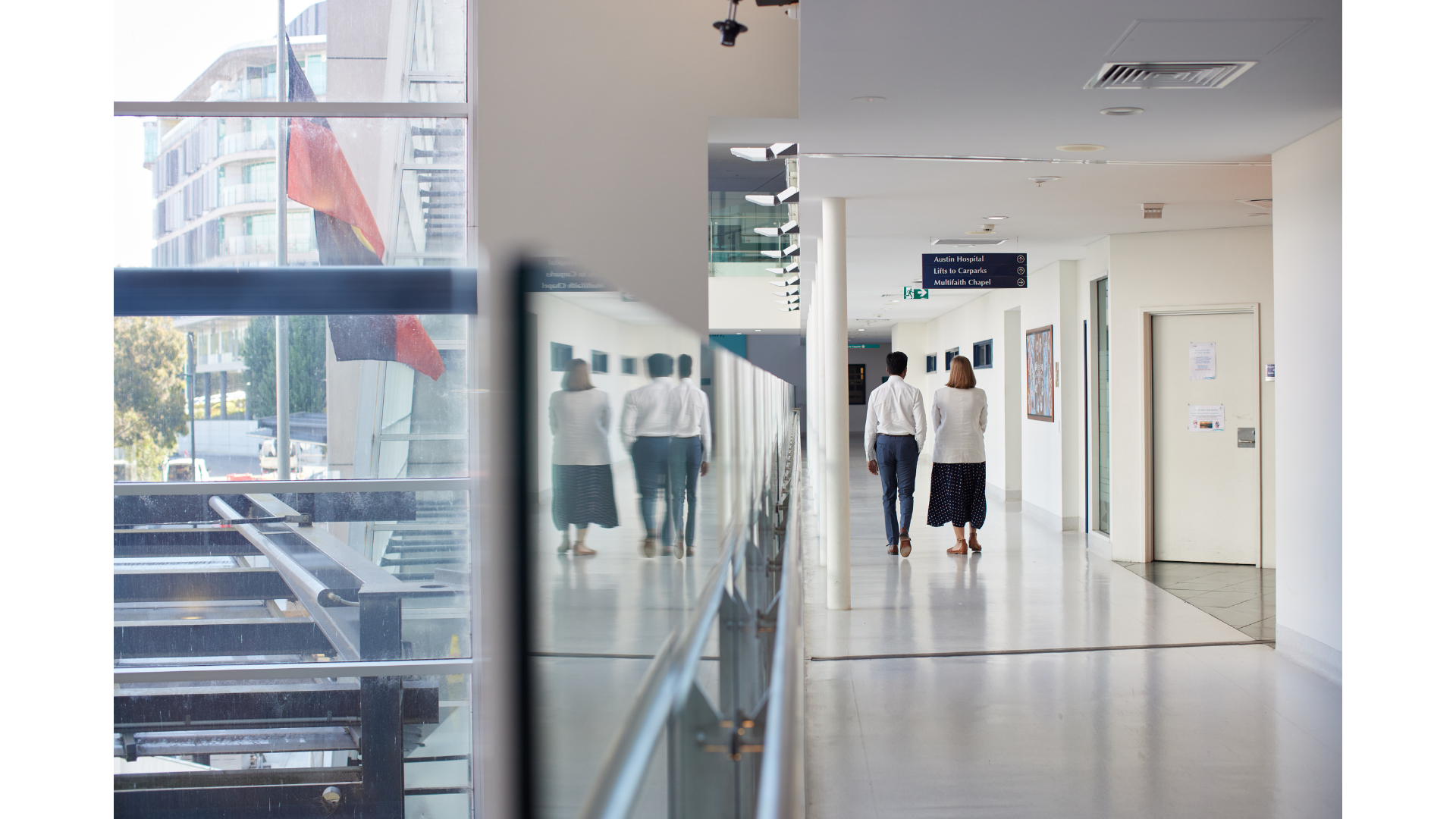 People walking down a hospital corridor with signs pointing to Austin Hospital, lifts to carparks, and a multifaith chapel.