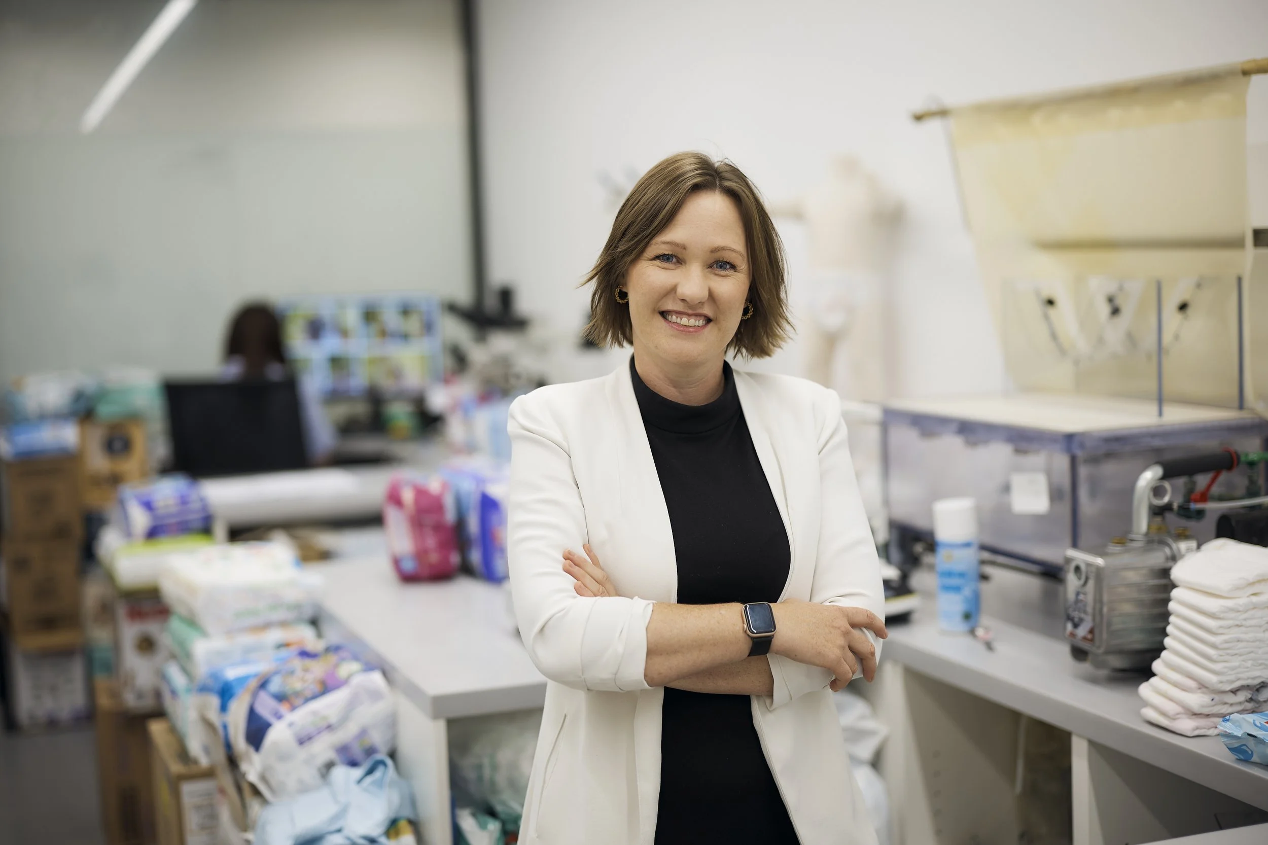 A woman standing in a laboratory or medical supply room with cross-arm posture, wearing a white lab coat and a smartwatch, smiling at the camera, surrounded by shelves filled with medical supplies and equipment.