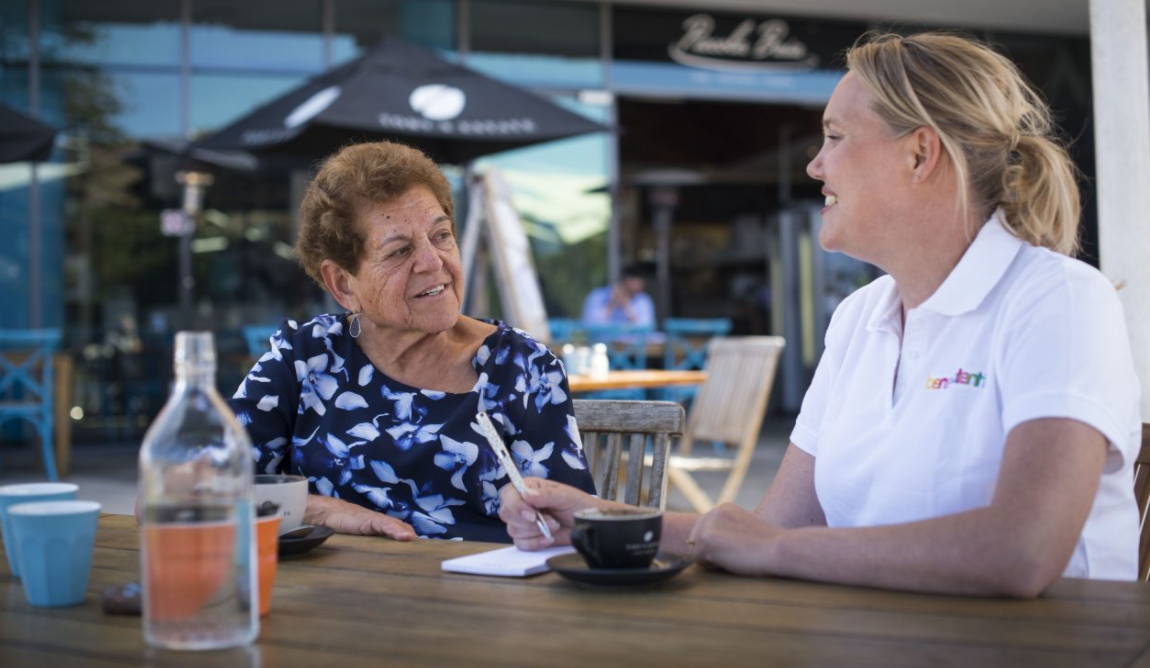 Two women sitting at a table outdoors, having a conversation, with a coffee cup, water bottle, and notebook on the table. One woman is elderly with short brown hair, wearing a blue and white patterned blouse, and the other is middle-aged with blonde hair tied back, wearing a white polo shirt with a rainbow-colored logo.