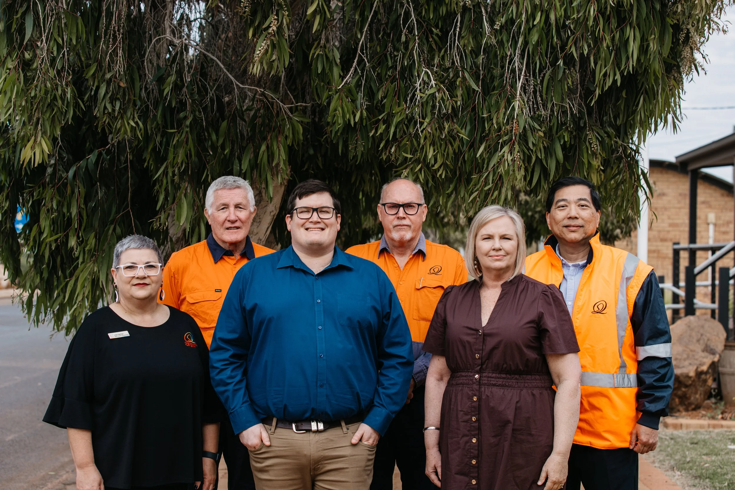 Group of six people standing outdoors in front of a large leafy tree, wearing casual and work clothing, smiling for the camera.