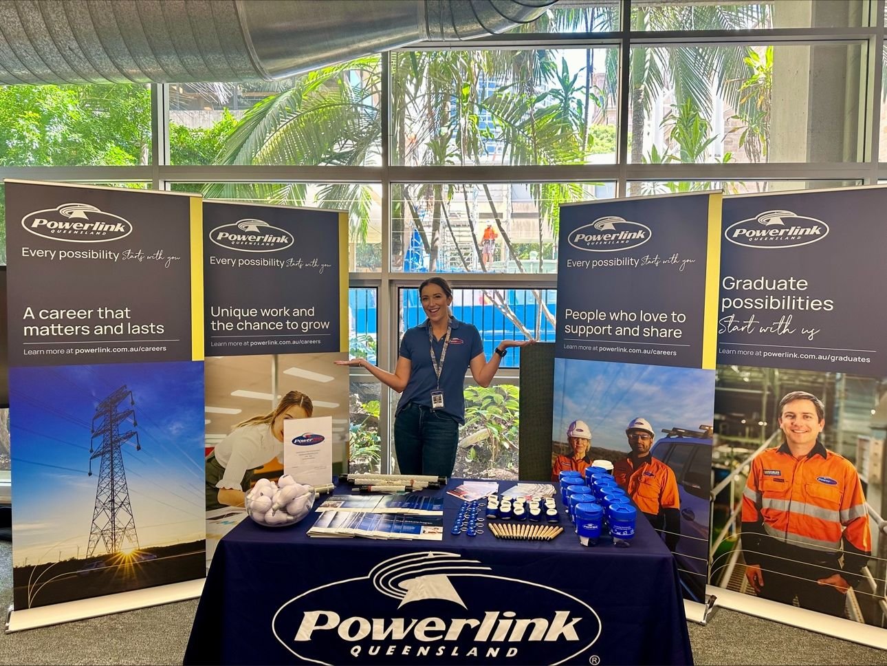 A woman at a Powerlink Queensland booth with promotional banners, tables with branded items, and a large window showing greenery outside.