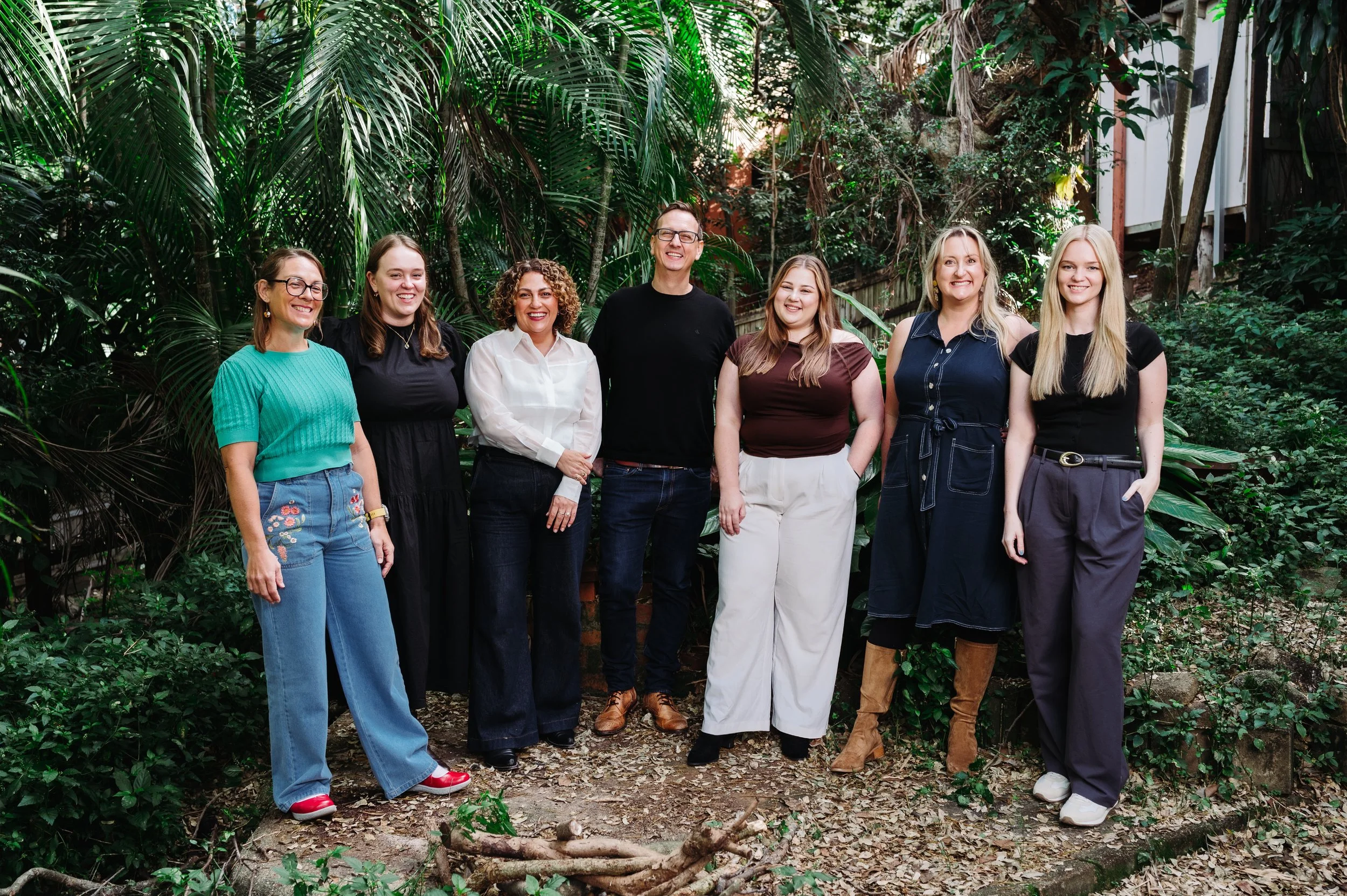 Group portrait of seven diverse adults standing outdoors in front of lush green plants, smiling and looking at the camera.