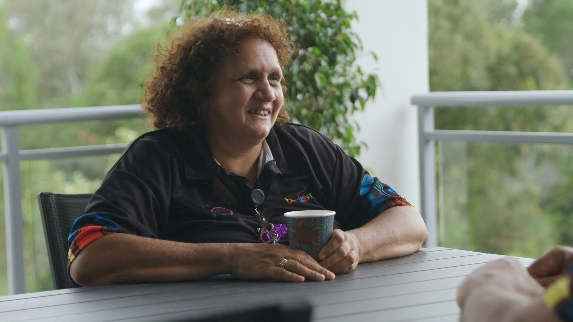 Woman sitting at an outdoor table, smiling and holding a colorful cup, with trees and a balcony railing in the background.