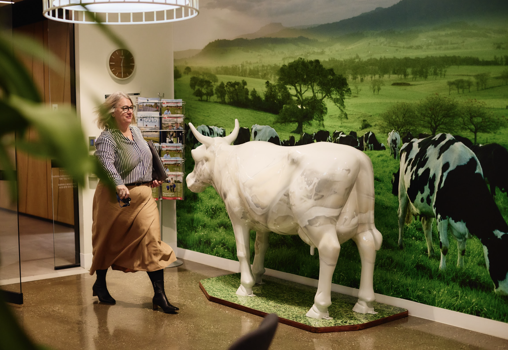 A woman with gray hair, glasses, and a striped shirt walking past a cow sculpture in front of a scenic countryside mural background.