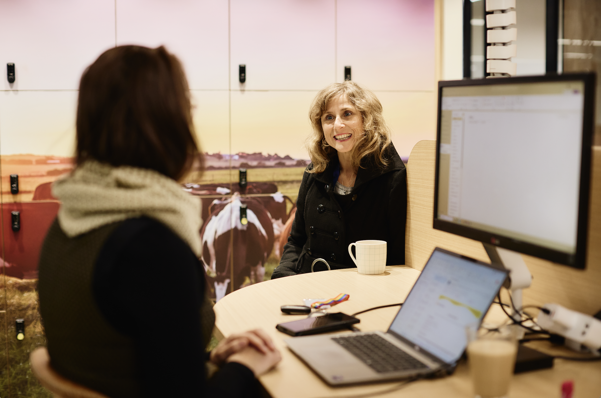 Two women having a conversation in an office with computers and a coffee mug on the table. The background features lockers and a landscape poster.