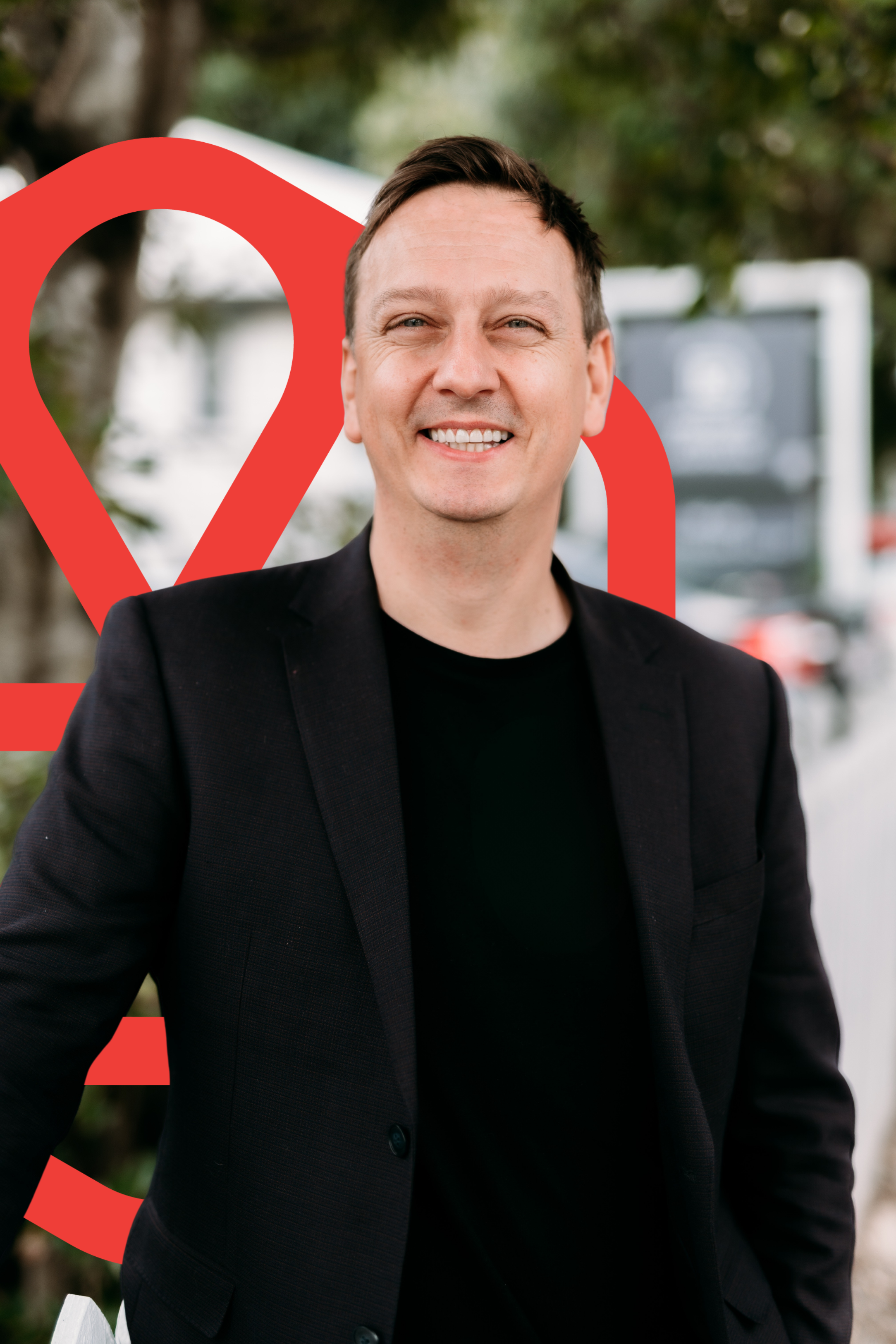 Smiling man wearing black blazer and black shirt standing outdoors with a tree and a blurry sign in the background.