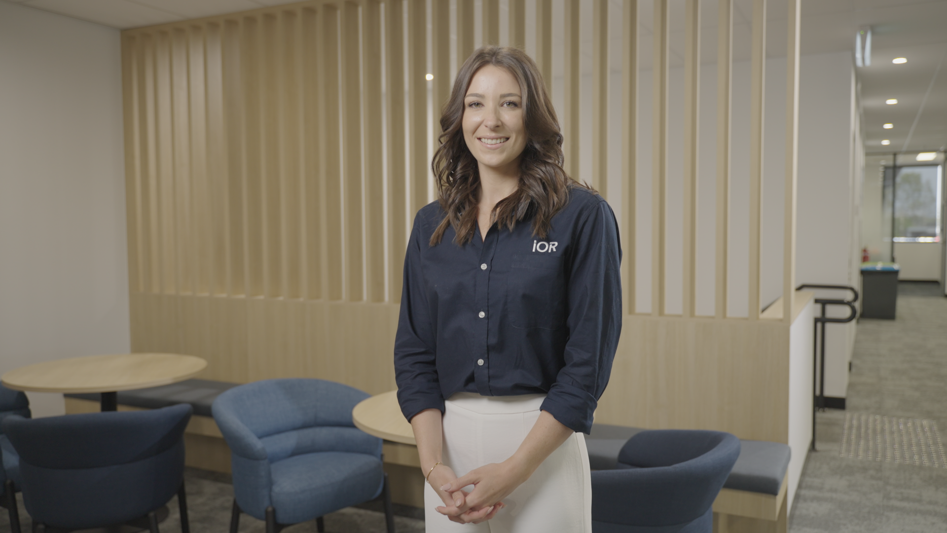 A smiling young woman with wavy brown hair, wearing a navy blue shirt with the logo 'IOR,' standing in a modern office lounge area with round tables and blue and black chairs, wooden slat wall panels, and large windows in the background.