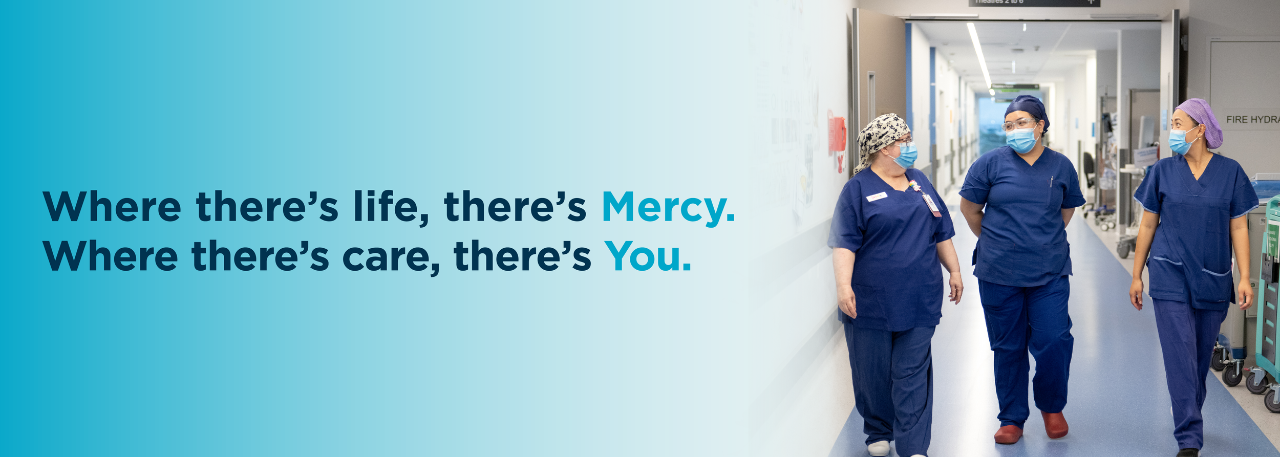 Three healthcare workers in blue scrubs and face masks walk down hospital corridor talking to each other.