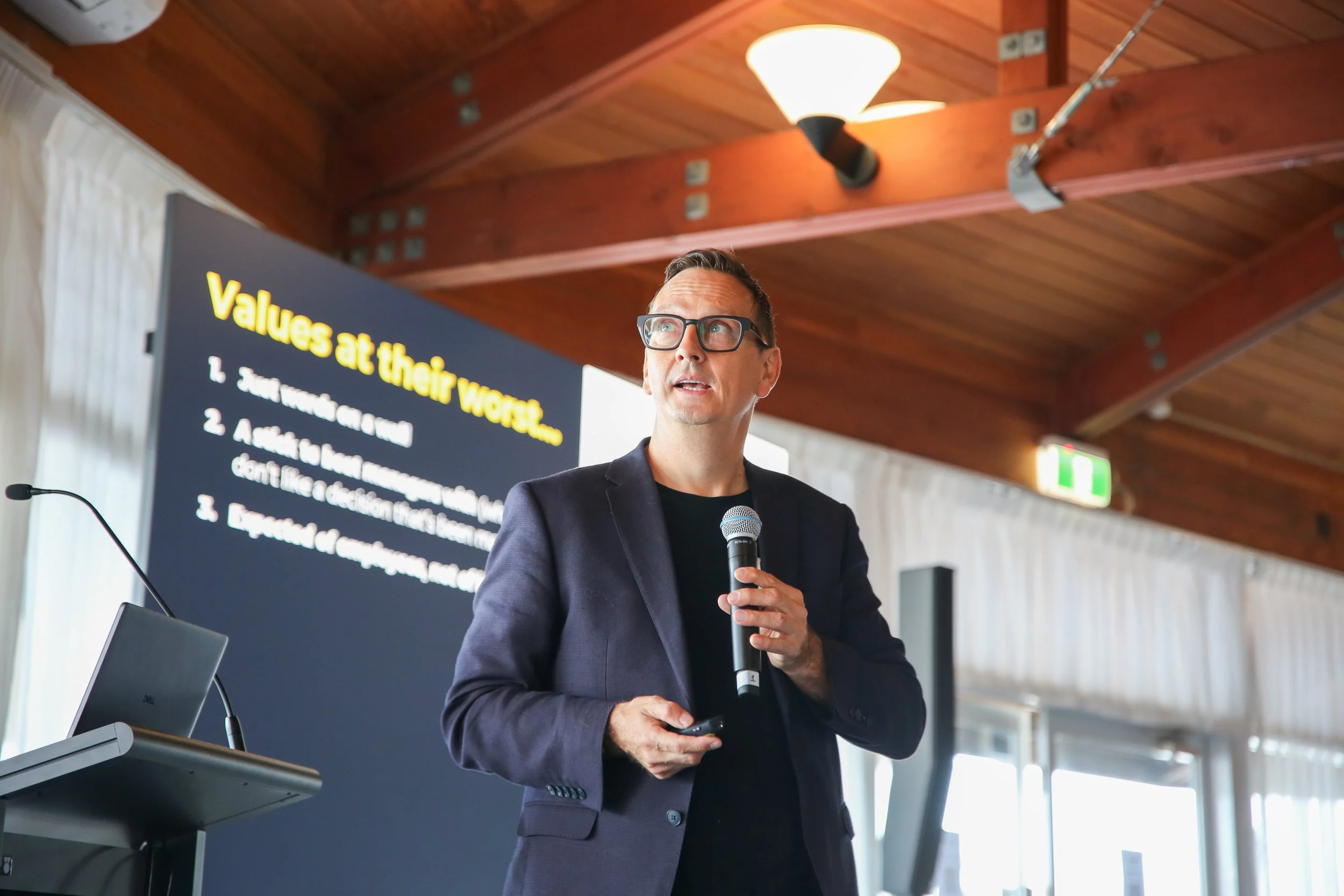 A man in a black blazer and glasses holding a microphone, giving a presentation in a room with wooden ceiling. A screen behind him displays text about values at work.