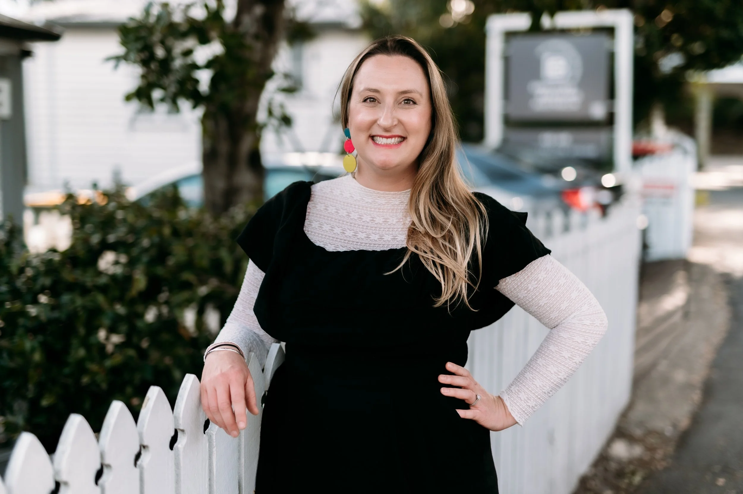 A woman with long blonde hair, smiling, standing outdoors by a white picket fence, wearing a black dress over a white lace top and colorful earrings.