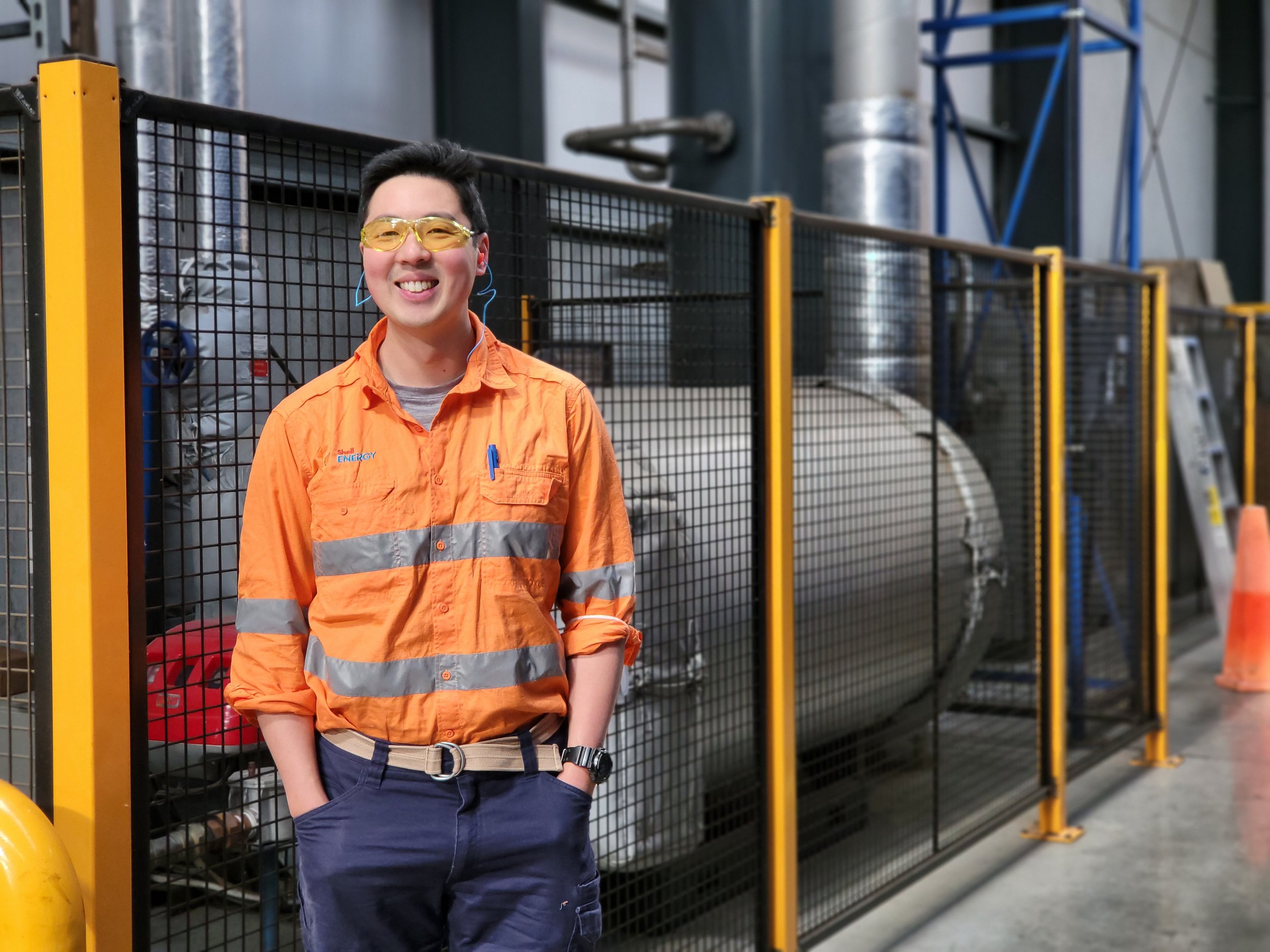 A smiling man wearing yellow safety goggles, an orange safety jacket with reflective stripes, and navy blue pants, stands with his hands in his pockets in an industrial or factory setting, with metal pipes and equipment in the background.