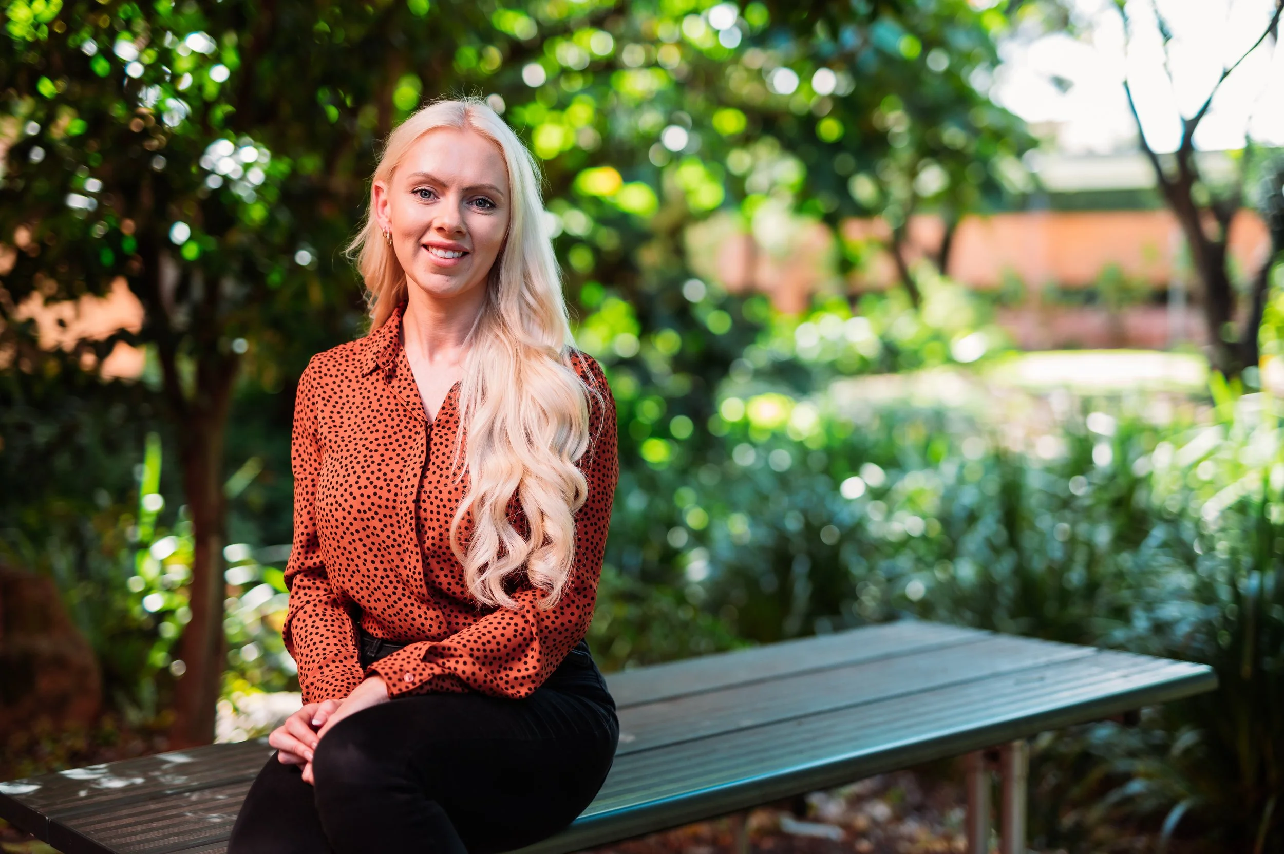 A woman with long blonde hair sitting on a park bench, smiling, with trees and greenery in the background.