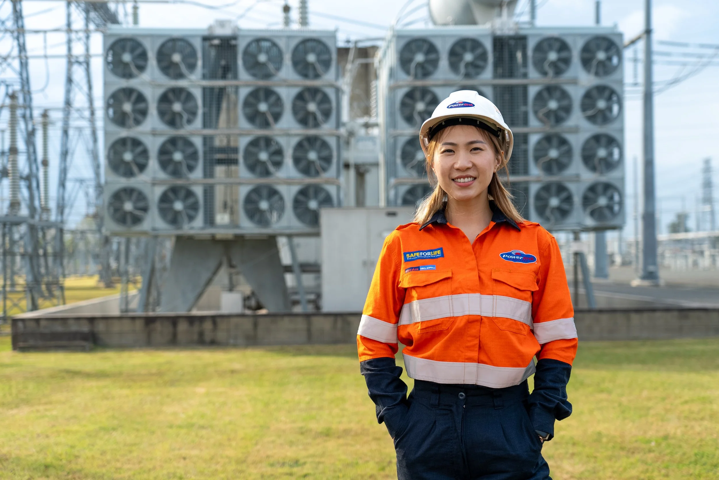A smiling woman wearing safety gear, including a white helmet and an orange safety vest, stands outdoors with power equipment and electrical infrastructure in the background.
