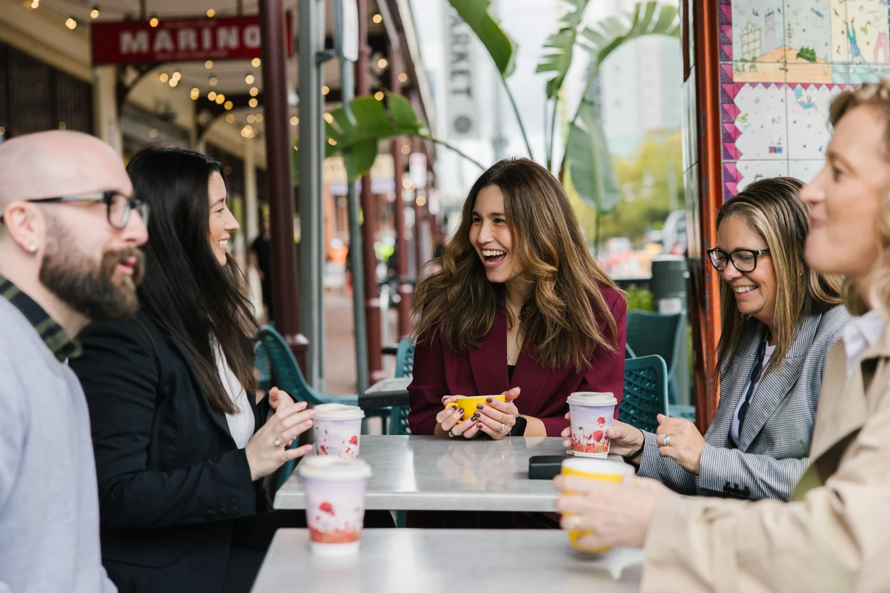 A group of six people sitting at a table outside a cafe, smiling and talking, with coffees in paper cups, on a sunny day with green plants and city buildings in the background.