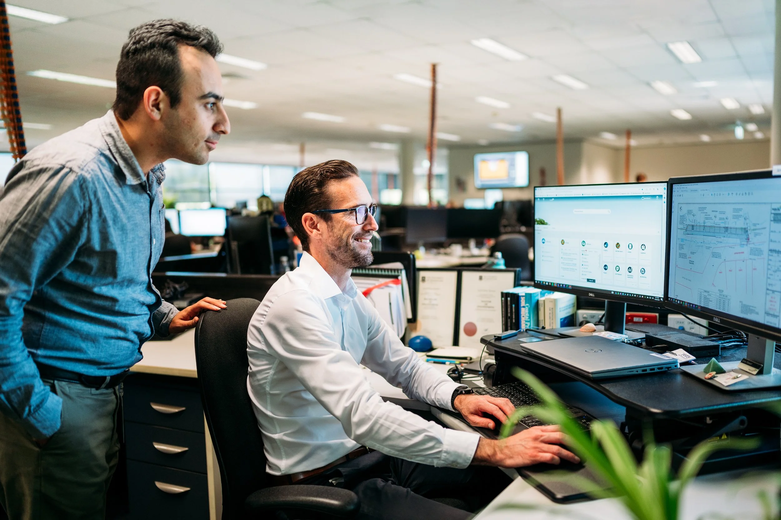 Two men working together at a computer in an office, one seated and the other standing, looking at the monitor displaying charts and data.